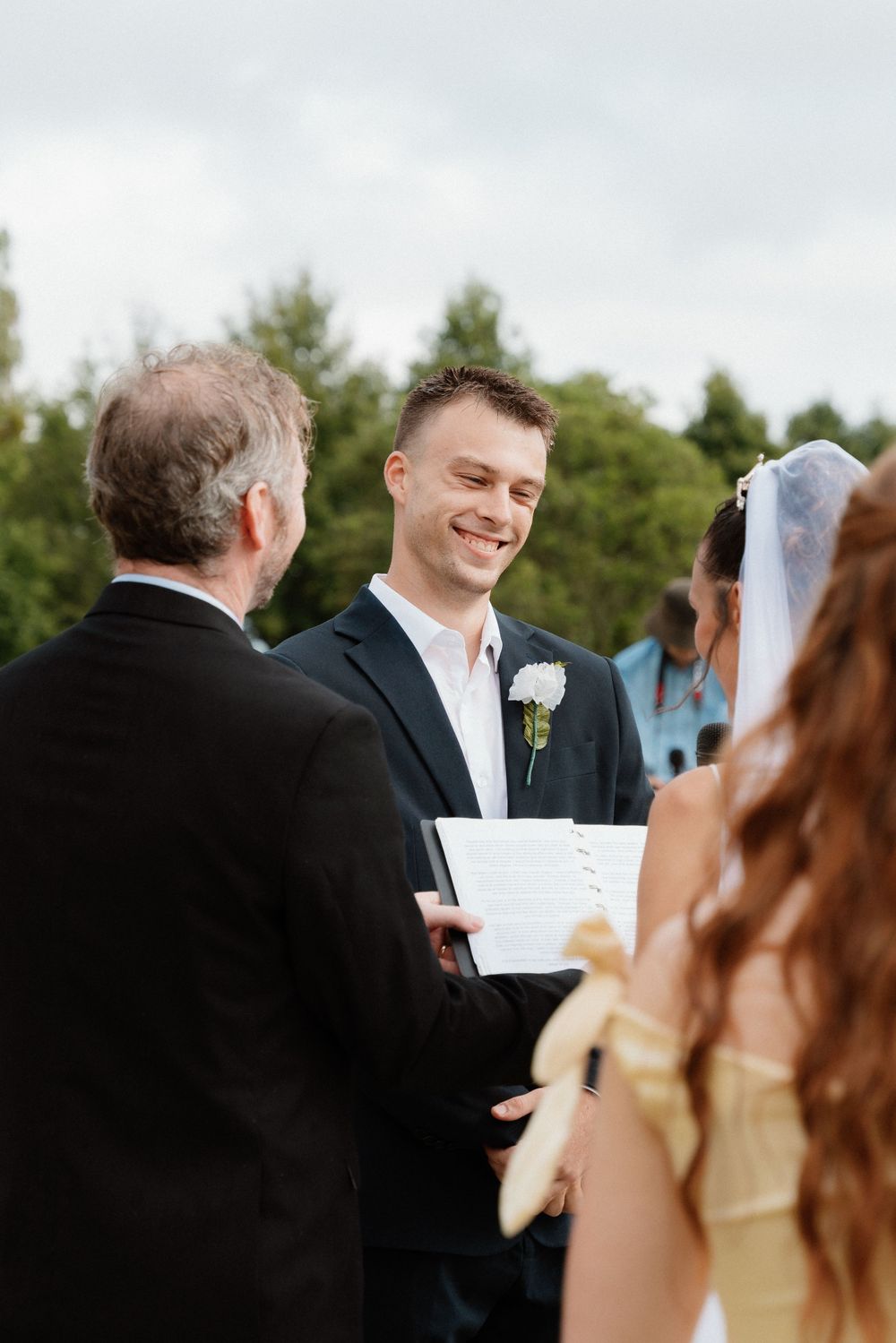 A wedding ceremony takes place outdoors as the officiant reads from papers while the couple exchanges their vows.