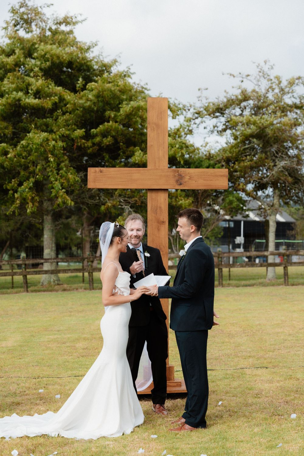 An outdoor wedding ceremony takes place in front of a wooden cross on a grassy lawn with trees in the background.