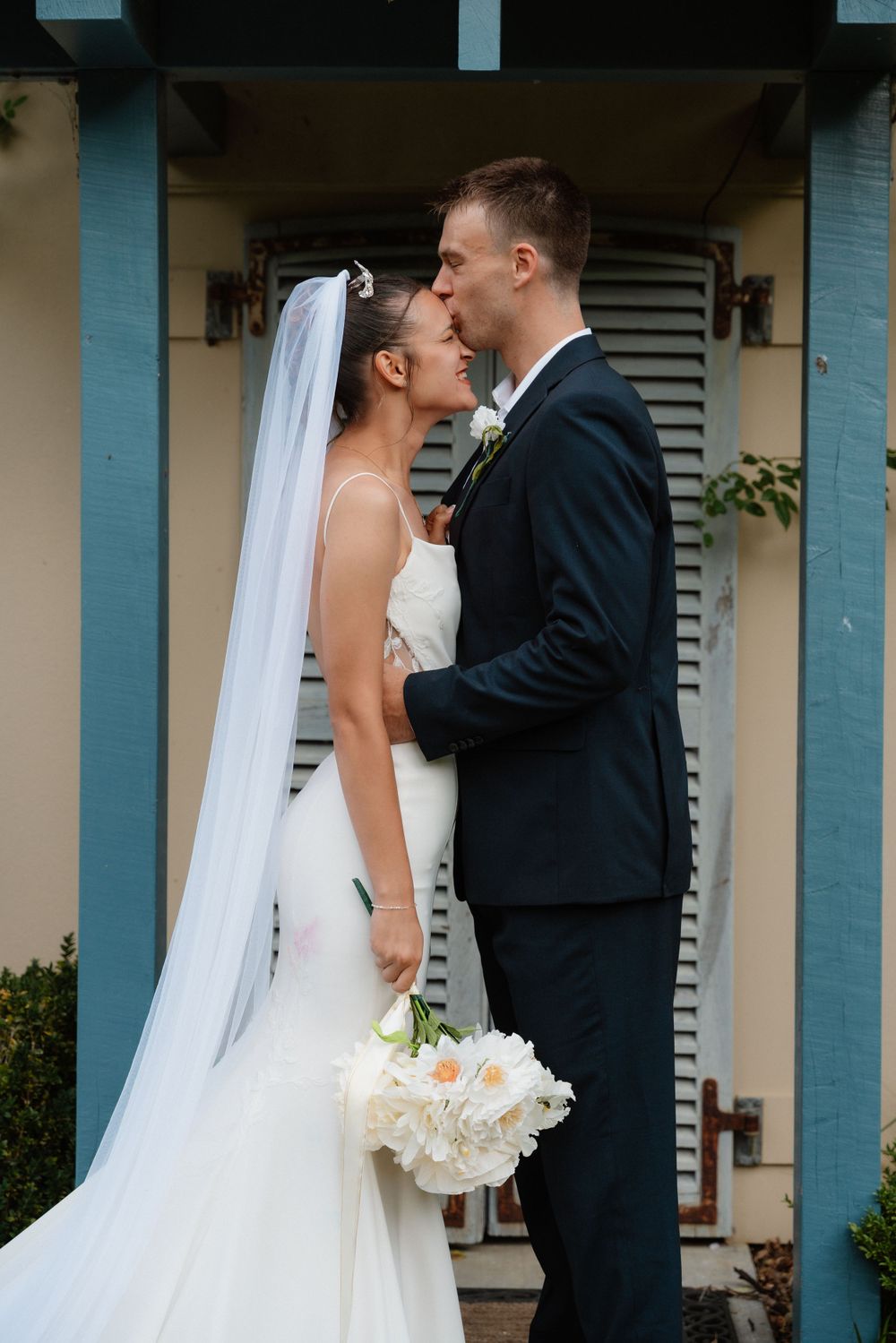 Newlywed couple shares intimate moment in front of turquoise pillars while bride holds white bouquet of flowers.