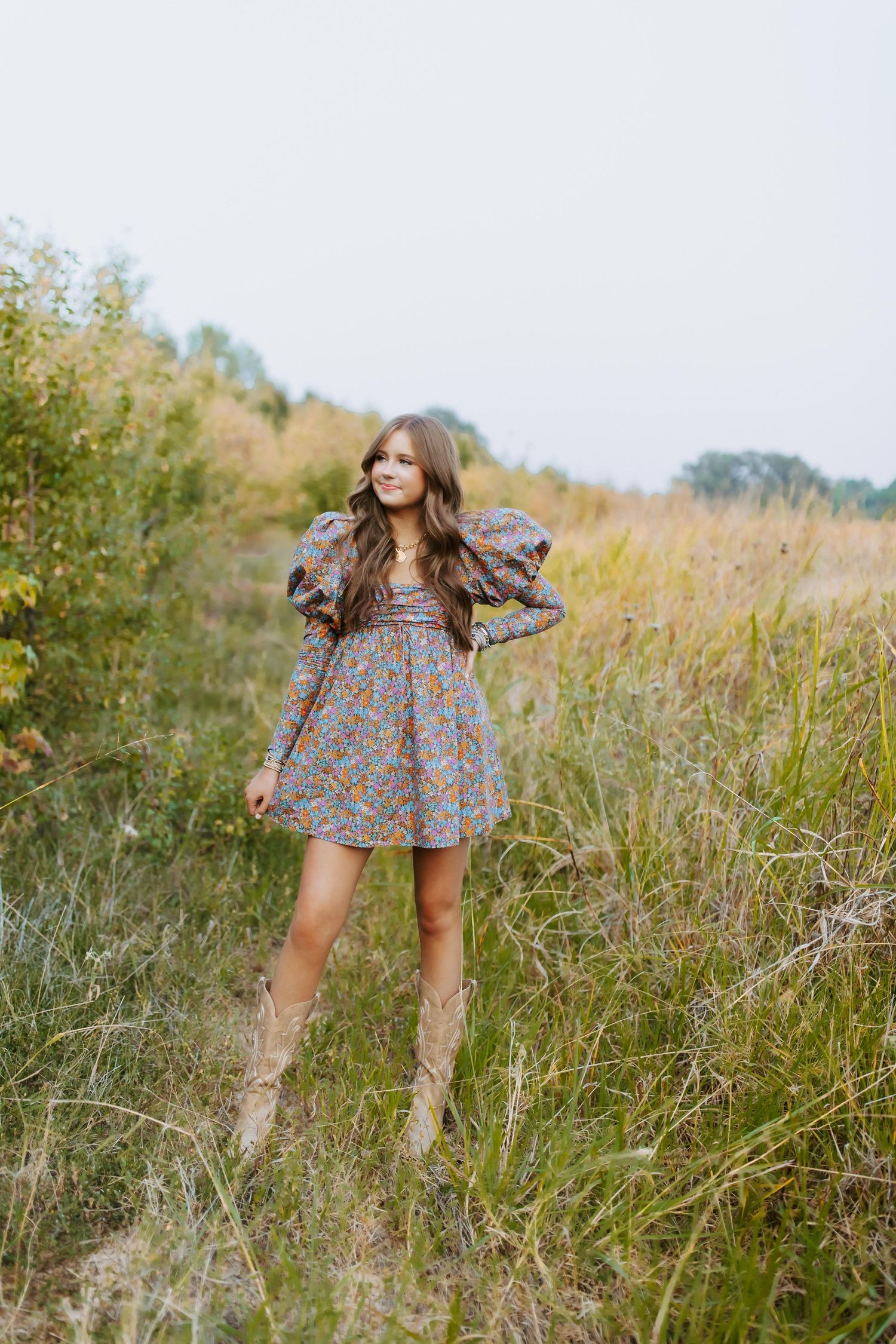 Person wearing a floral dress and tan boots poses in a rustic field with tall grass and autumn foliage.