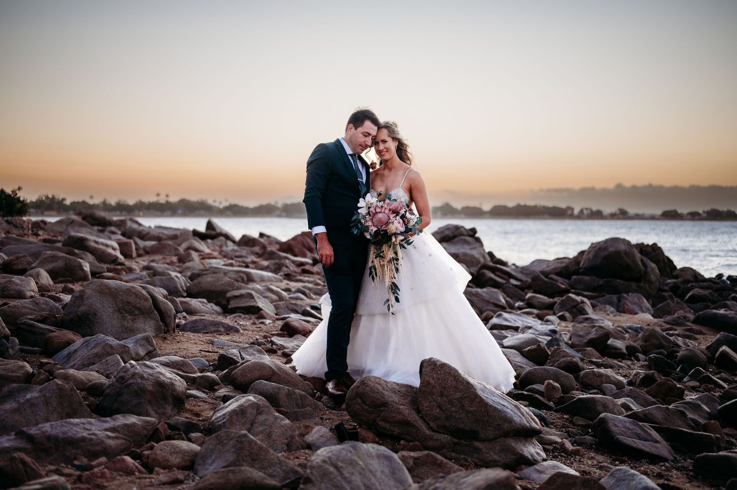 Romantic sunset wedding photo of a couple standing on rocky coastline.