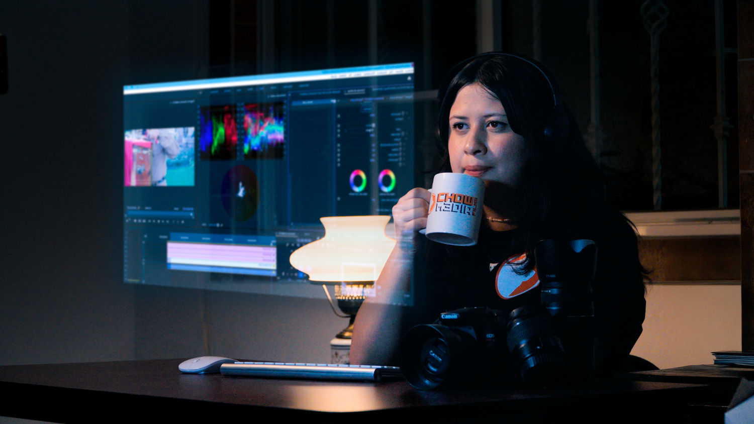 A woman holds a coffee mug while sitting at a desk with video editing software displayed on a monitor behind her.
