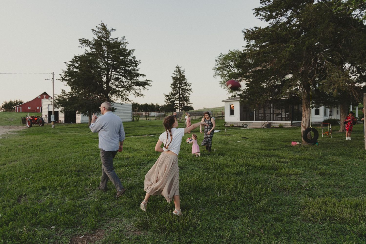 People enjoying a relaxed outdoor gathering on a grassy lawn with red barn buildings in the background at dusk.