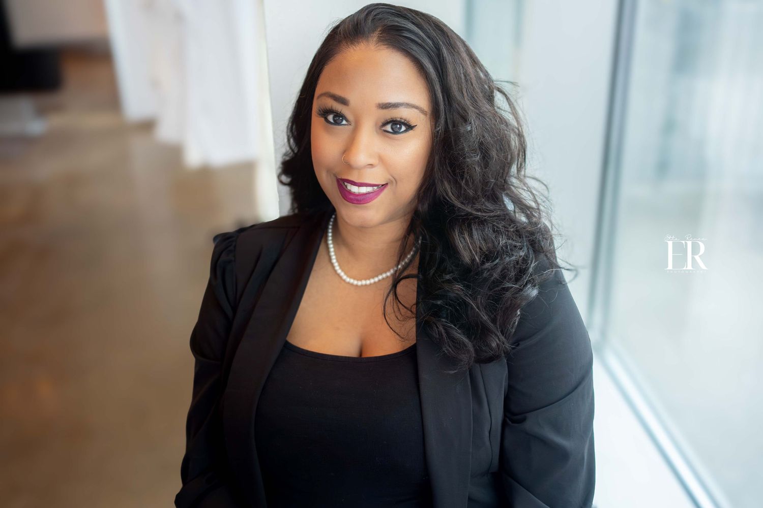 Professional business portrait of someone in a black blazer and top against a bright window background.