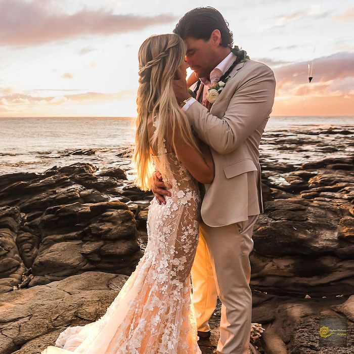 Couple shares romantic moment on rocky beach during golden hour sunset in Hawaii wearing formal wedding attire.