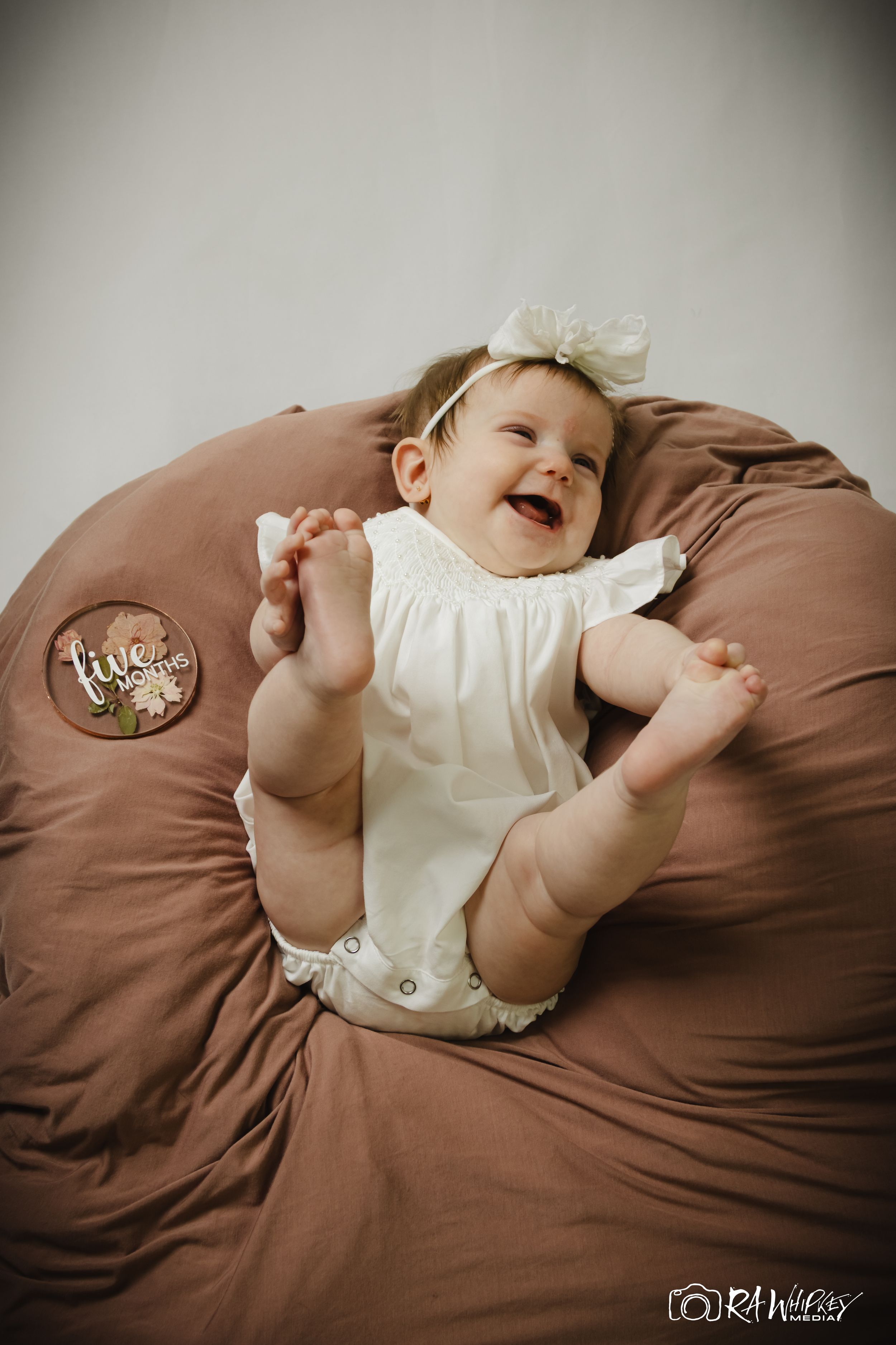 Laughing baby wearing white outfit lying on brown cushion.