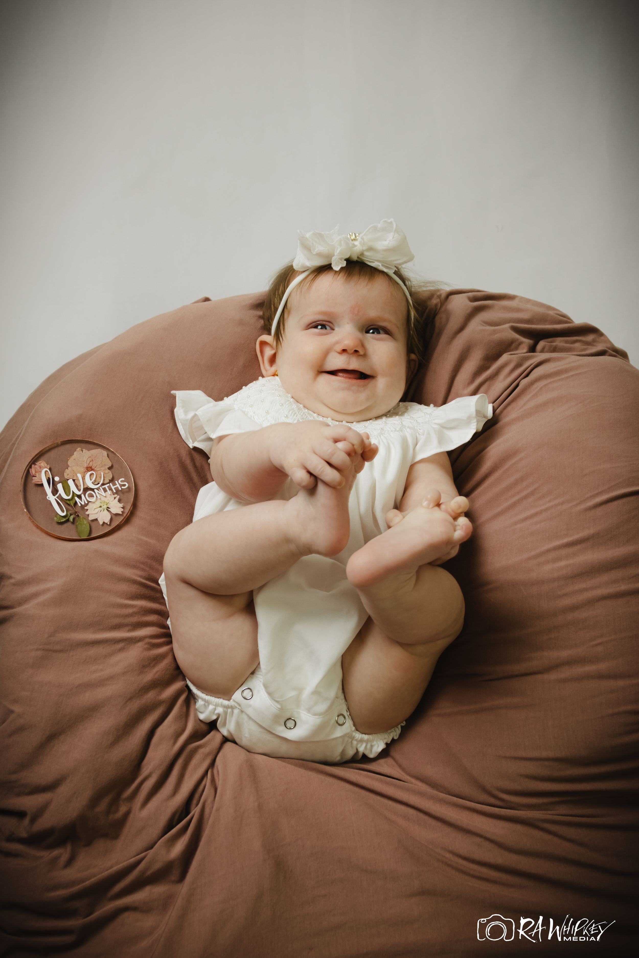 Baby in white outfit with bow smiles while lying on brown pillow holding their feet.