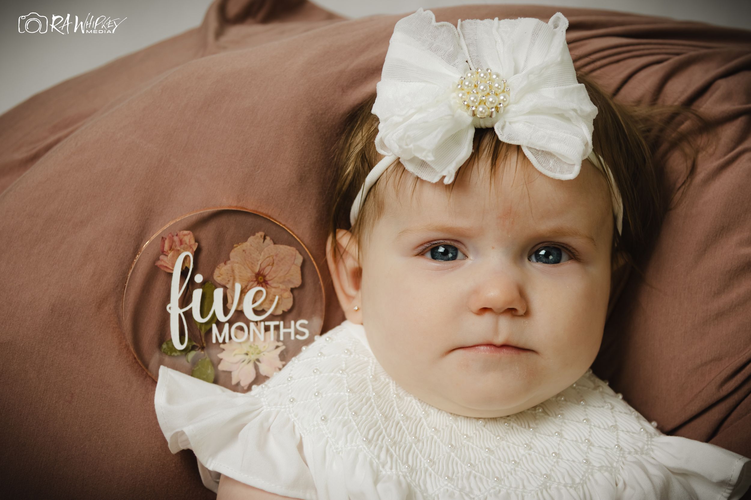 A baby wearing a white bow headband and dress lies on a brown cushion with a decorative wooden sign.