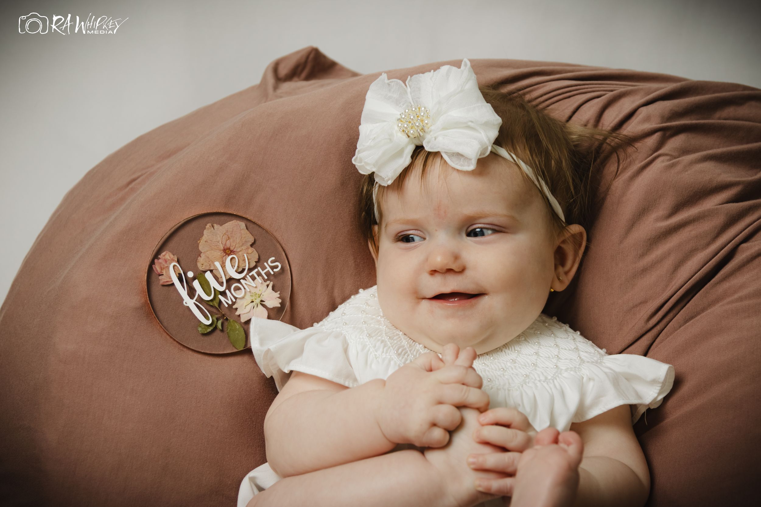 Smiling infant in white dress with bow headband reclines on brown cushion.