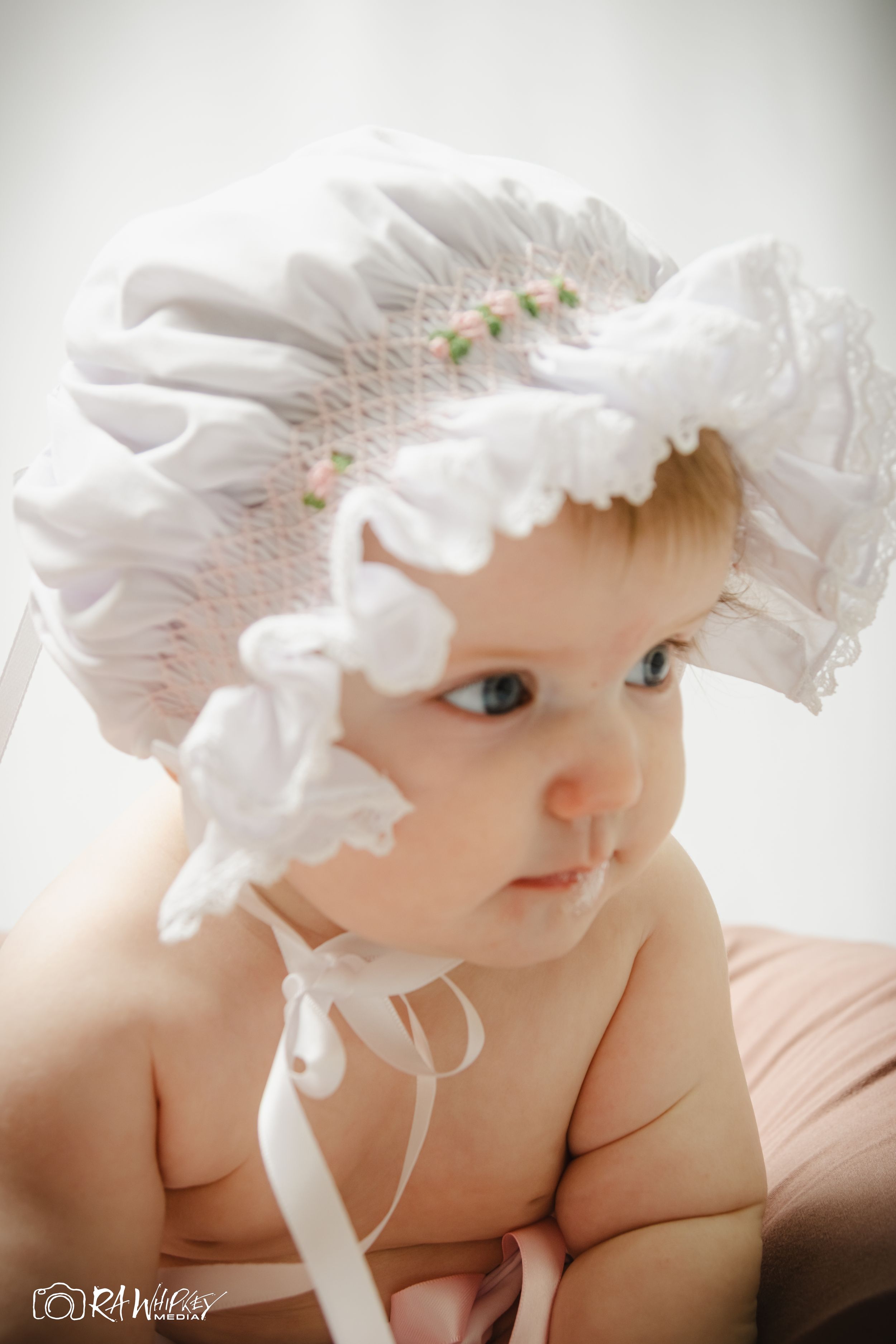 Baby wearing an ornate white bonnet with pink flowers looks to the side while lying down.