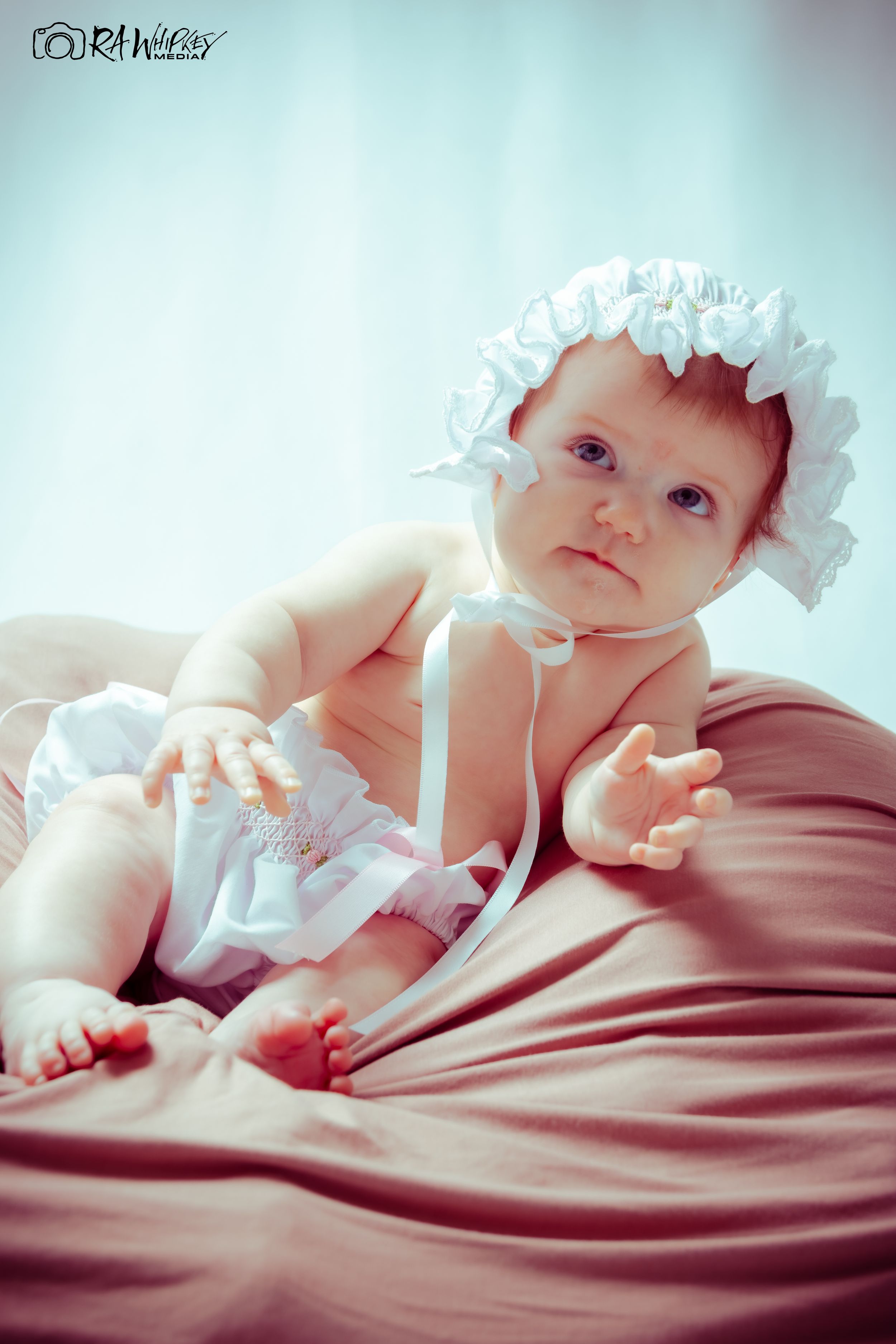 Baby in white ruffled bonnet lies on mauve fabric looking at camera.