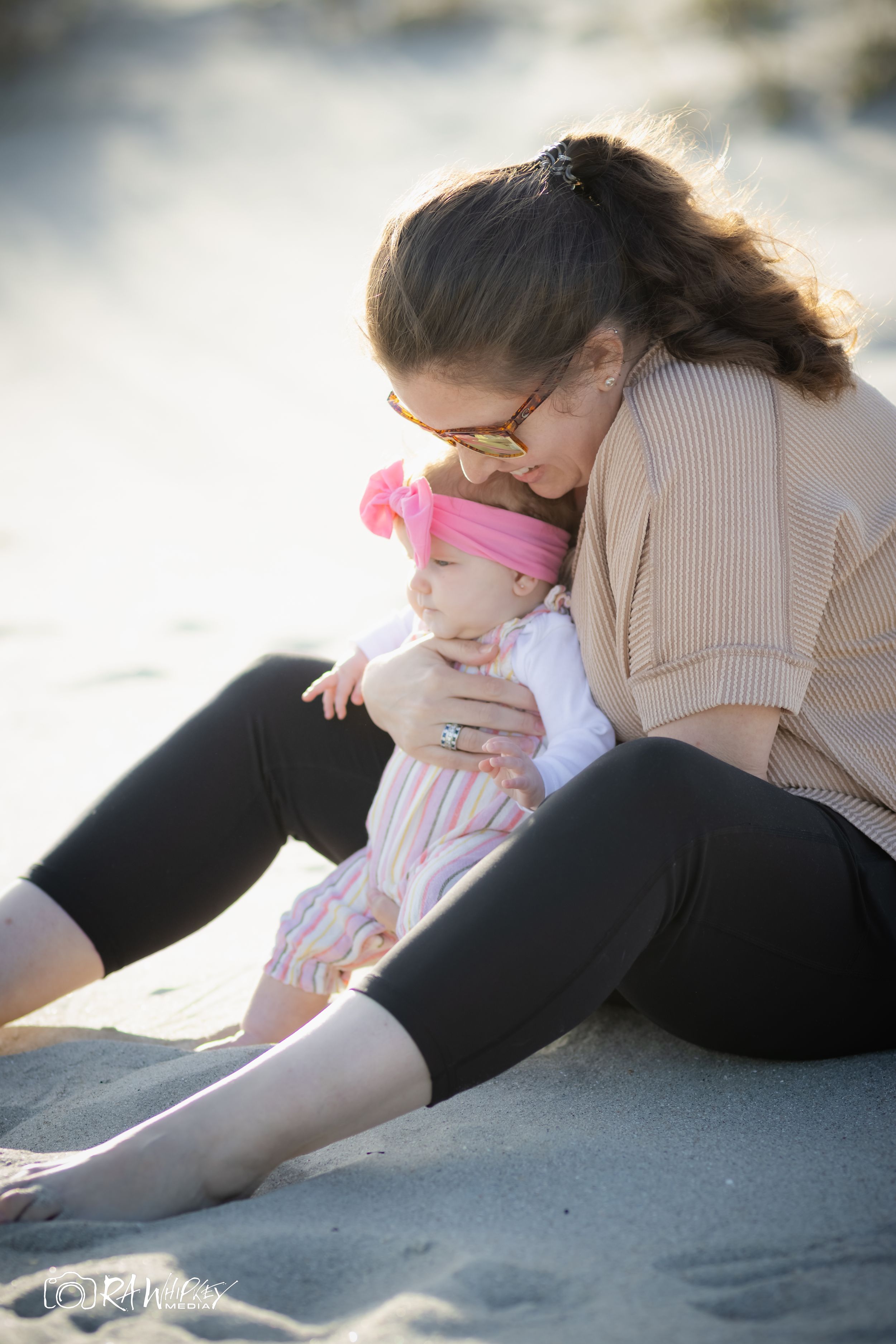 Mother and baby share tender moment while sitting on beach sand at sunset.