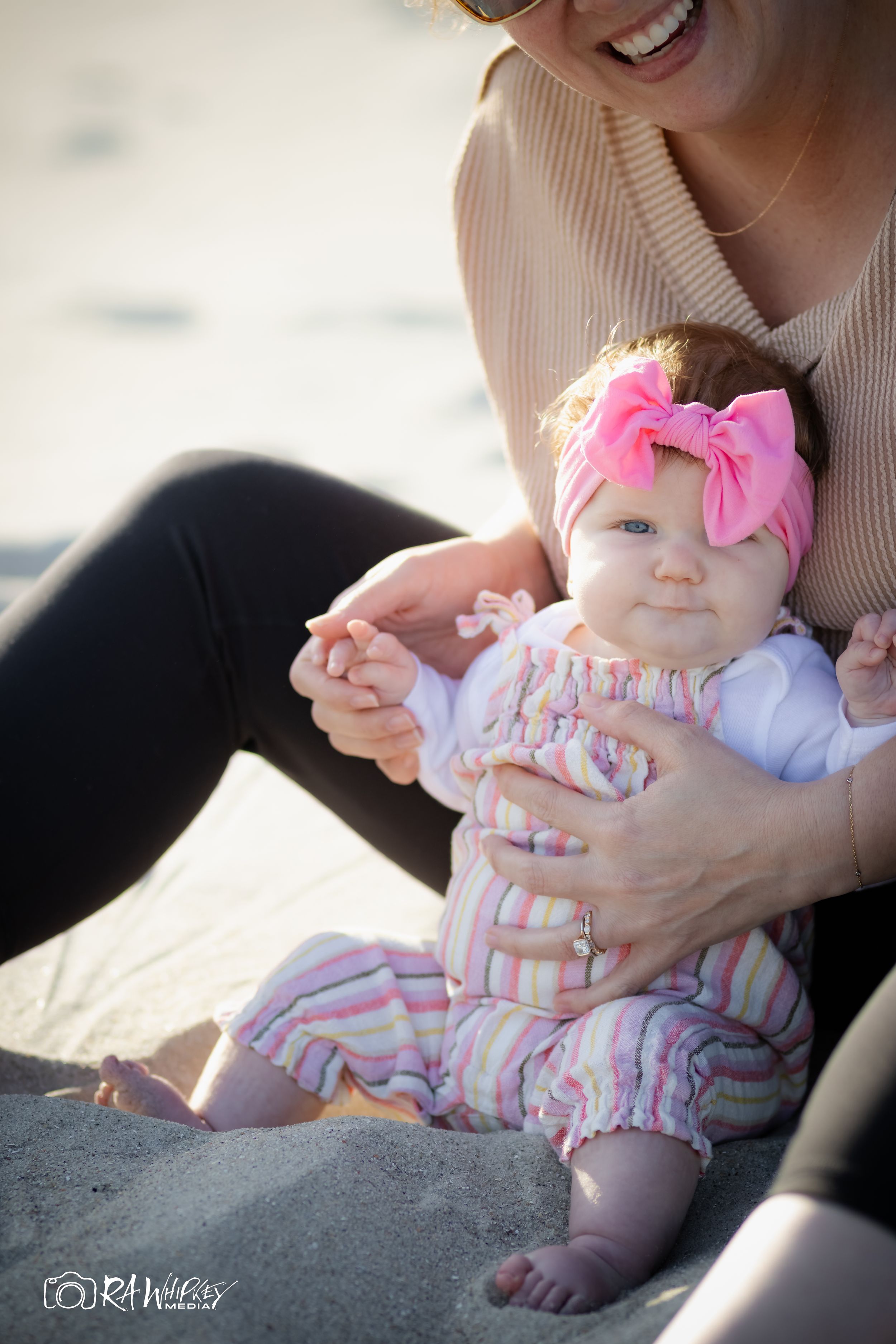 Baby in striped outfit with pink bow sits on beach with caregiver.