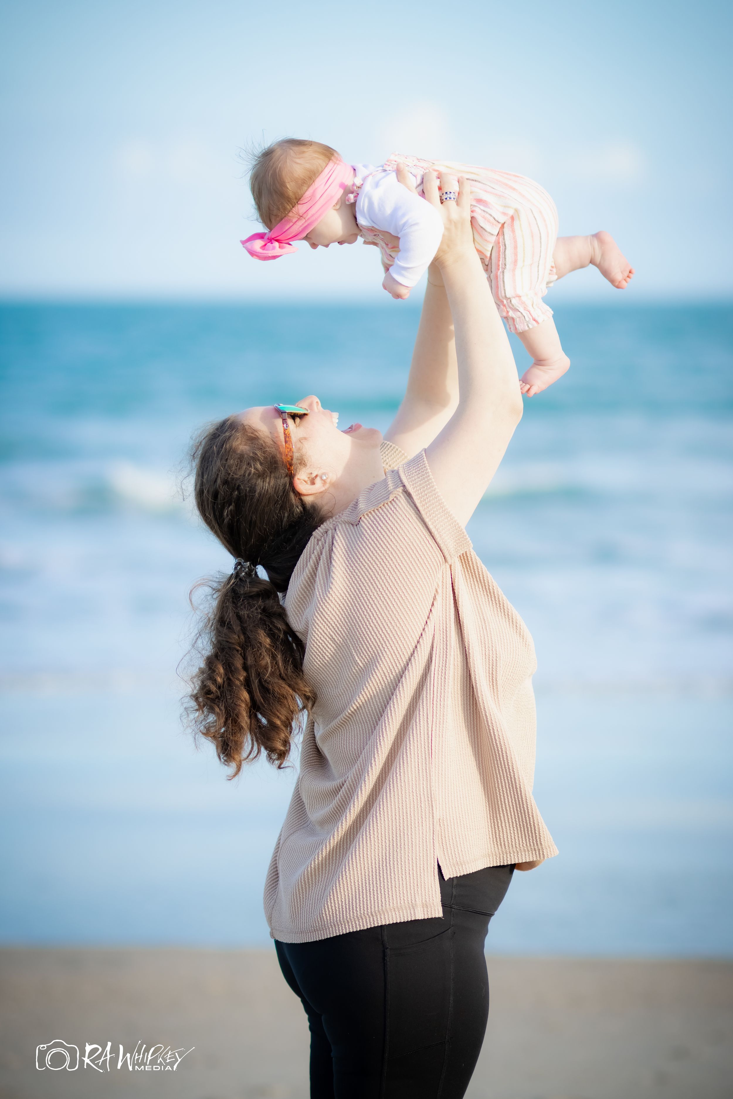 Parent lifts baby wearing pink bow against ocean backdrop at beach.