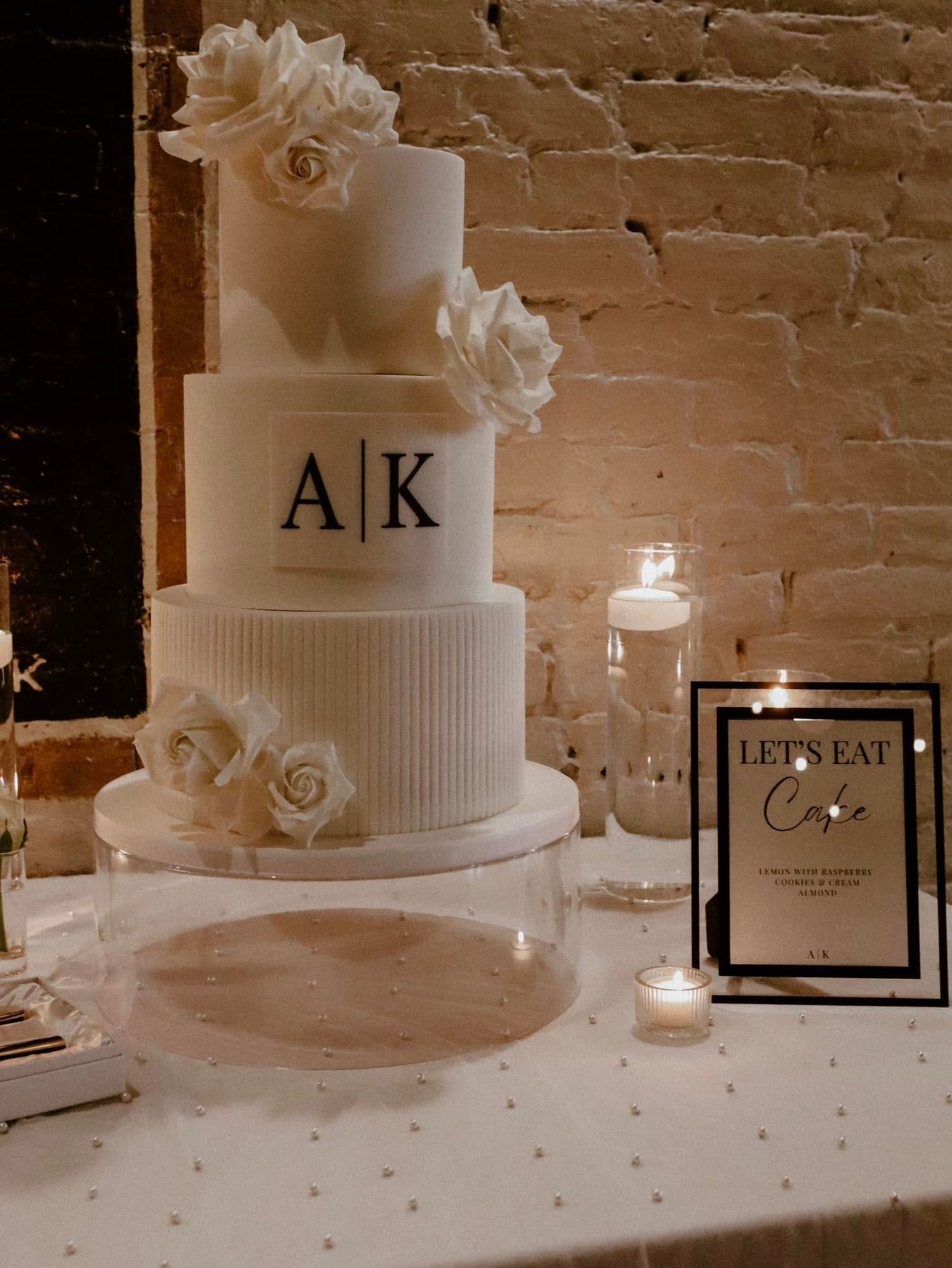 Elegant white wedding cake with initials A K decorated with white roses and placed on table with candles and brick wall backdrop.