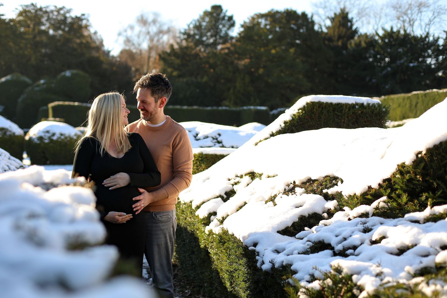 Two people stand together in a snow-covered garden with sunlight streaming through trees in the background.