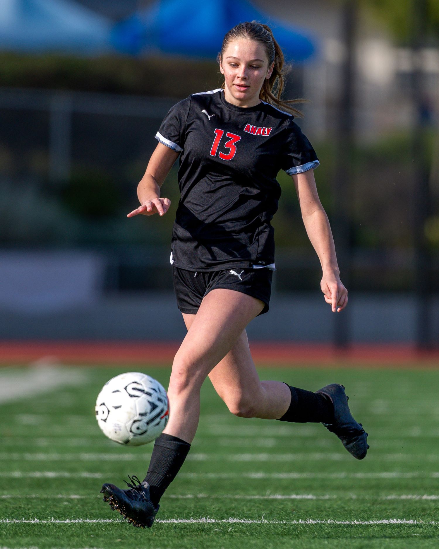 Soccer player in black uniform kicks ball during outdoor match on artificial turf.