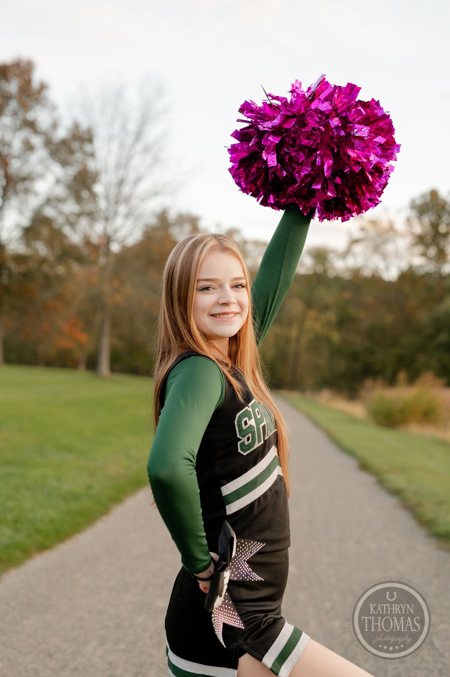 Cheerleader in green and black uniform holds up bright pink pom pom on park path.