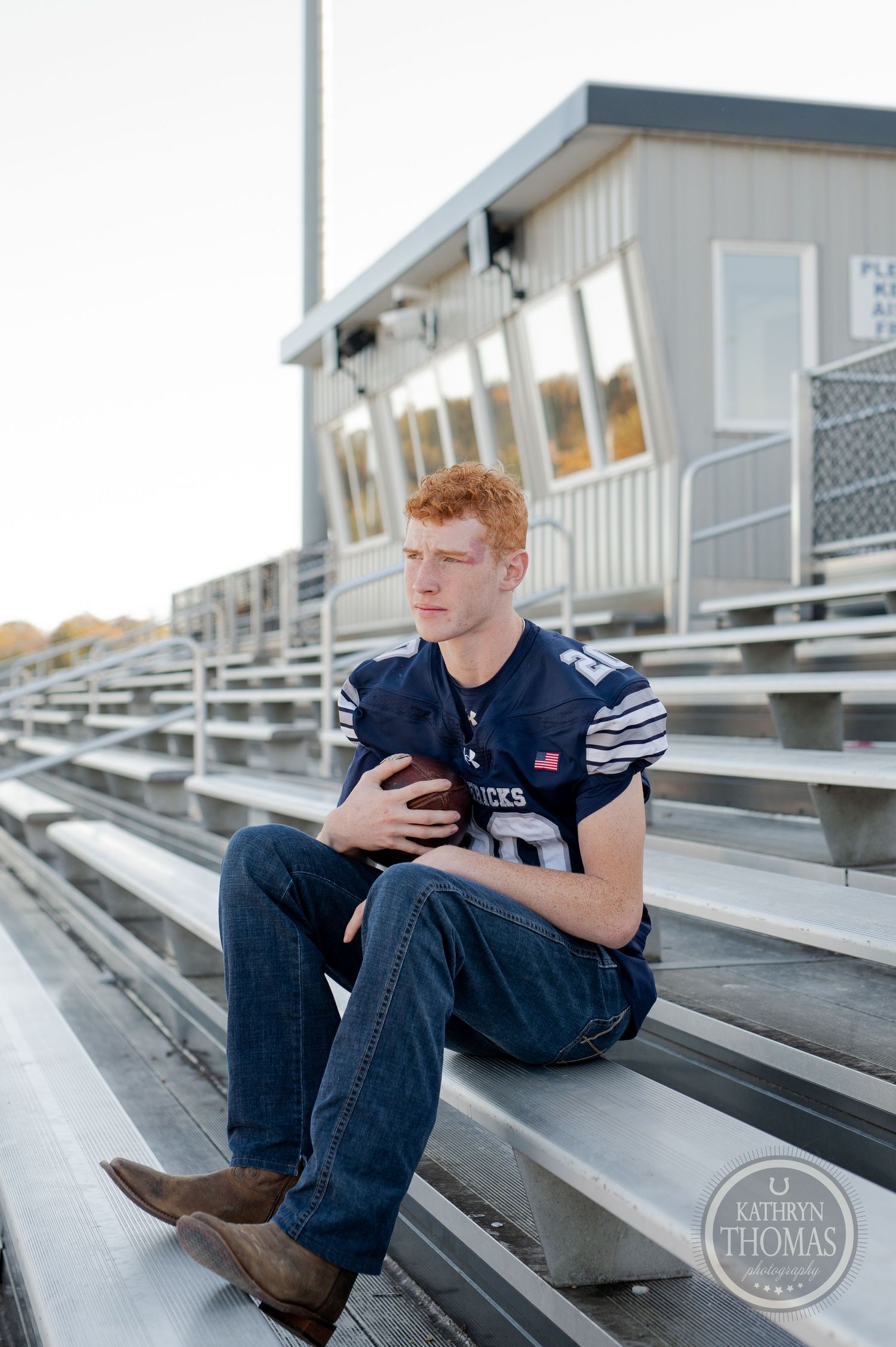 Football player in navy jersey sits on metal bleachers near press box.