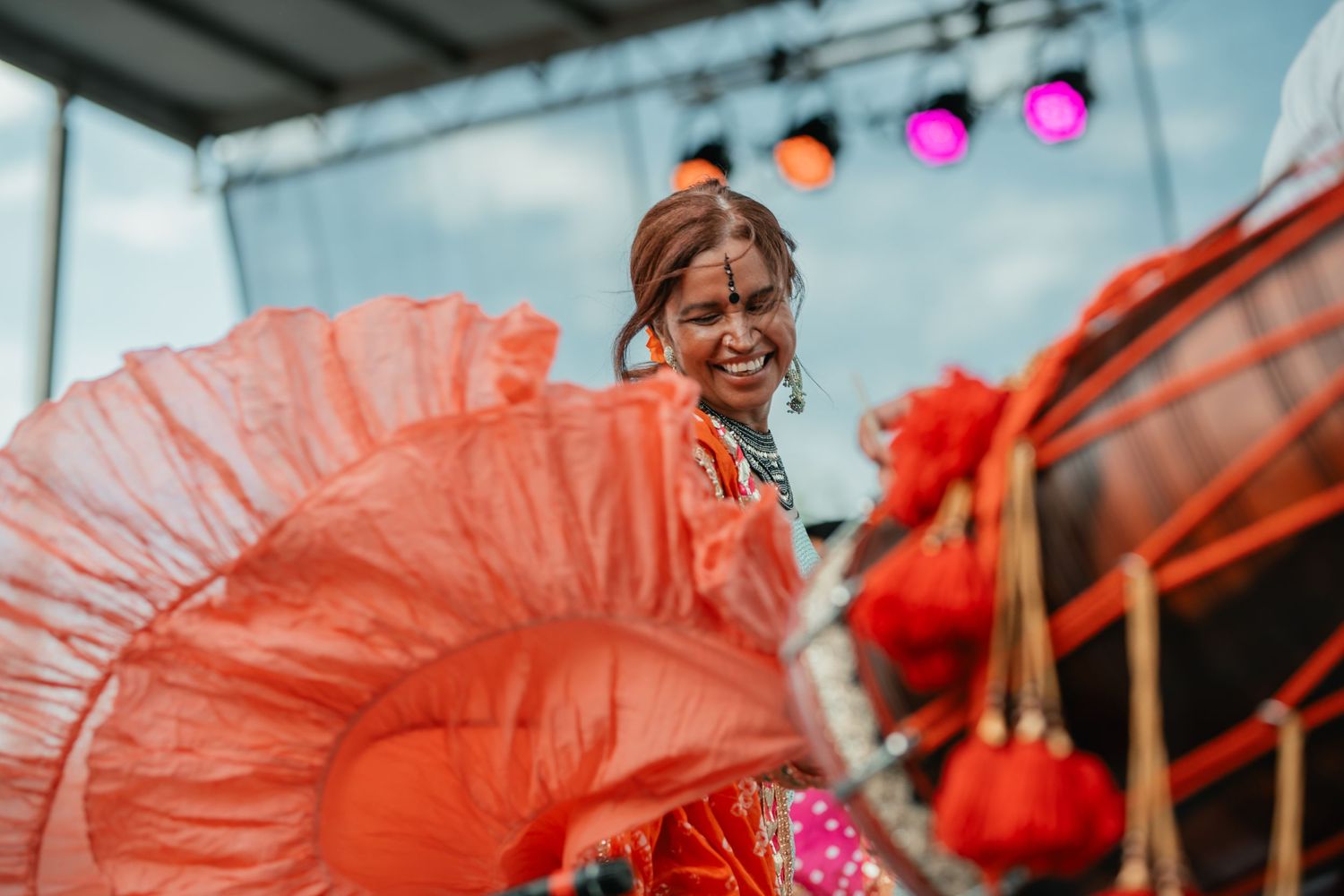 Performer in vibrant orange ruffled costume dancing on outdoor stage with colorful lights.