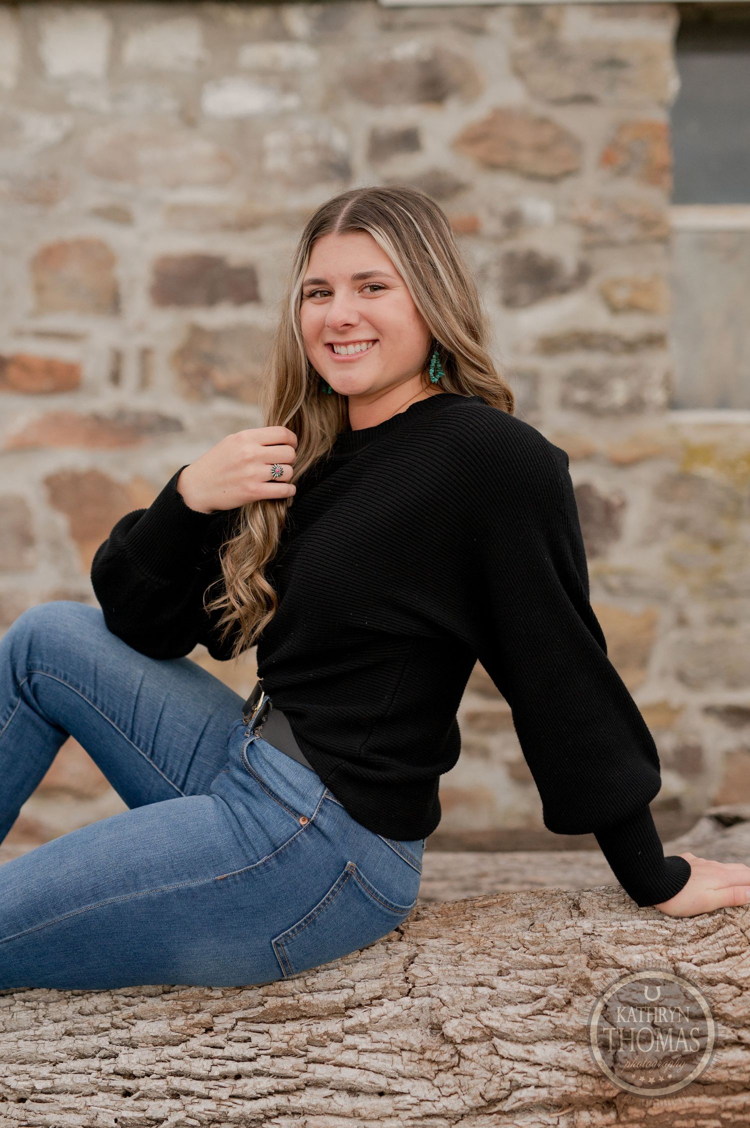 Casual portrait on stone wall wearing black sweater and blue jeans in outdoor setting.