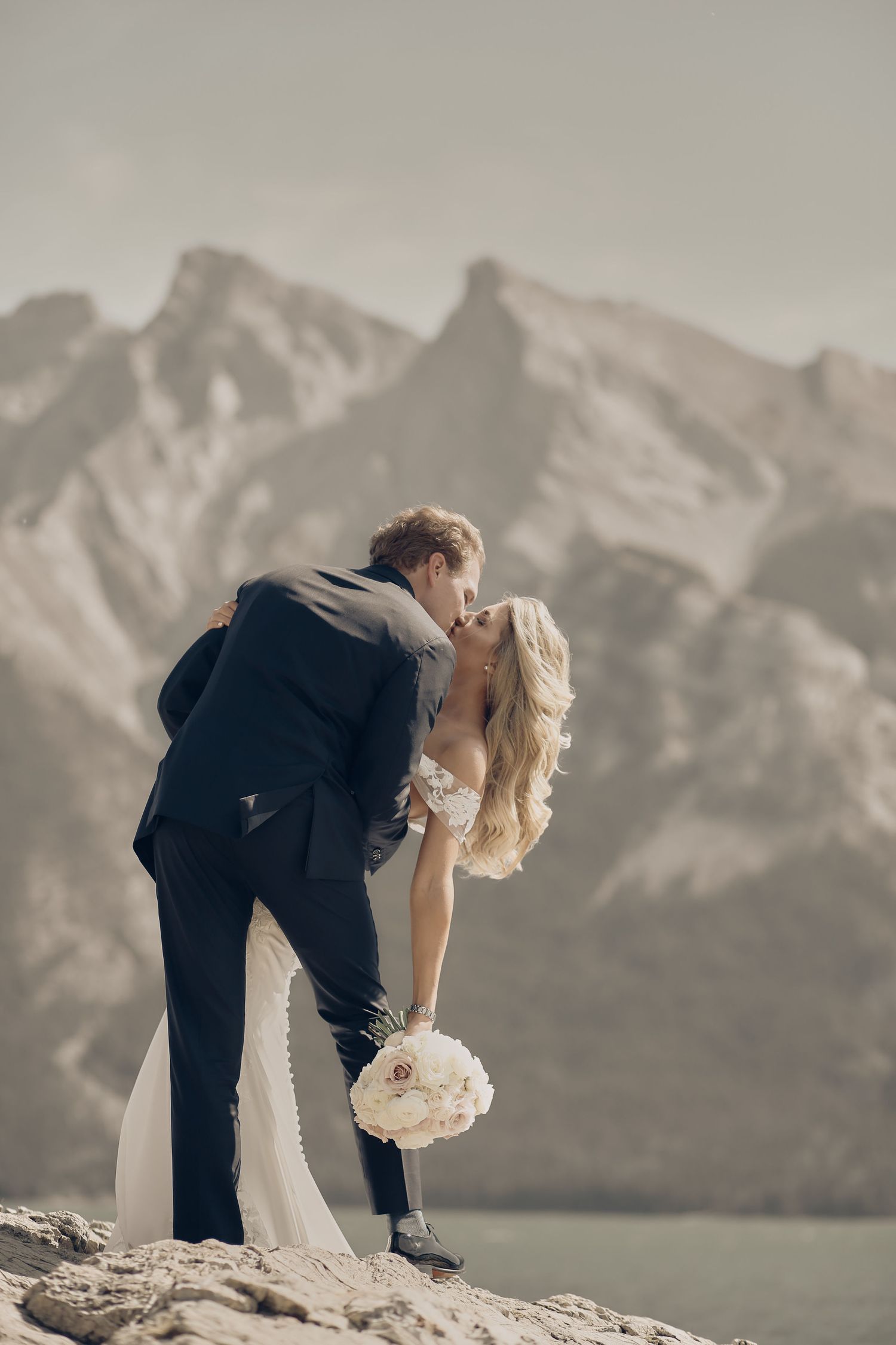 A romantic wedding couple shares a kiss on a rocky outcrop with mountain peaks and a lake in the background on a helicopter photo shoot