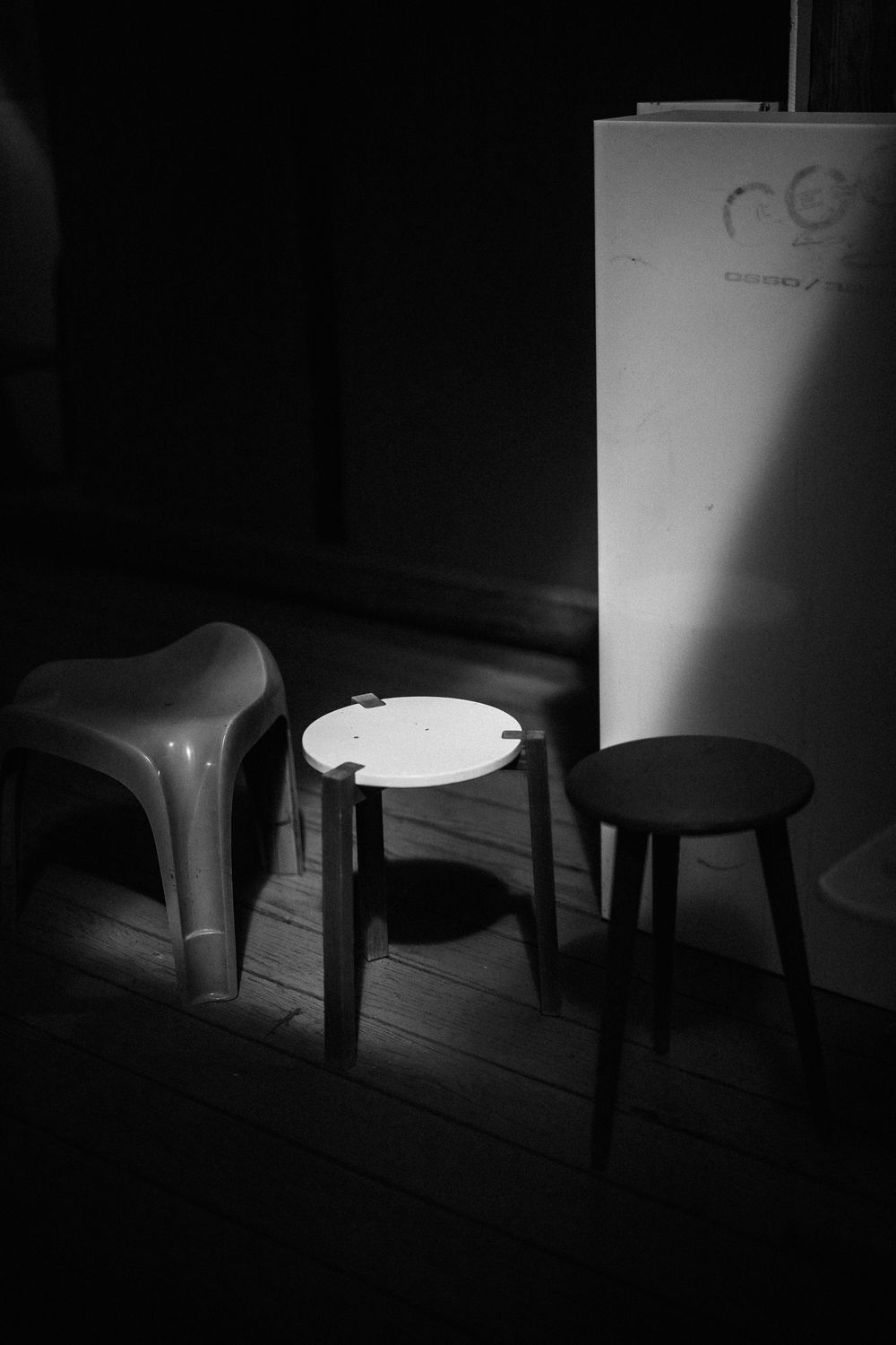 Black and white photo of three chairs in dim lighting - a plastic chair and two small stools on wooden floorboards.