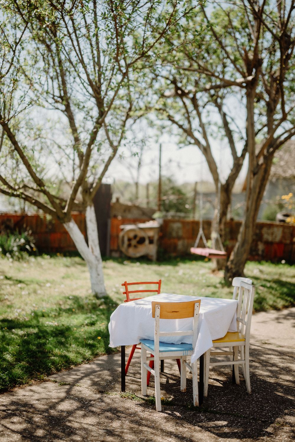 Outdoor dining table with white tablecloth set under blooming trees in sunlit garden.