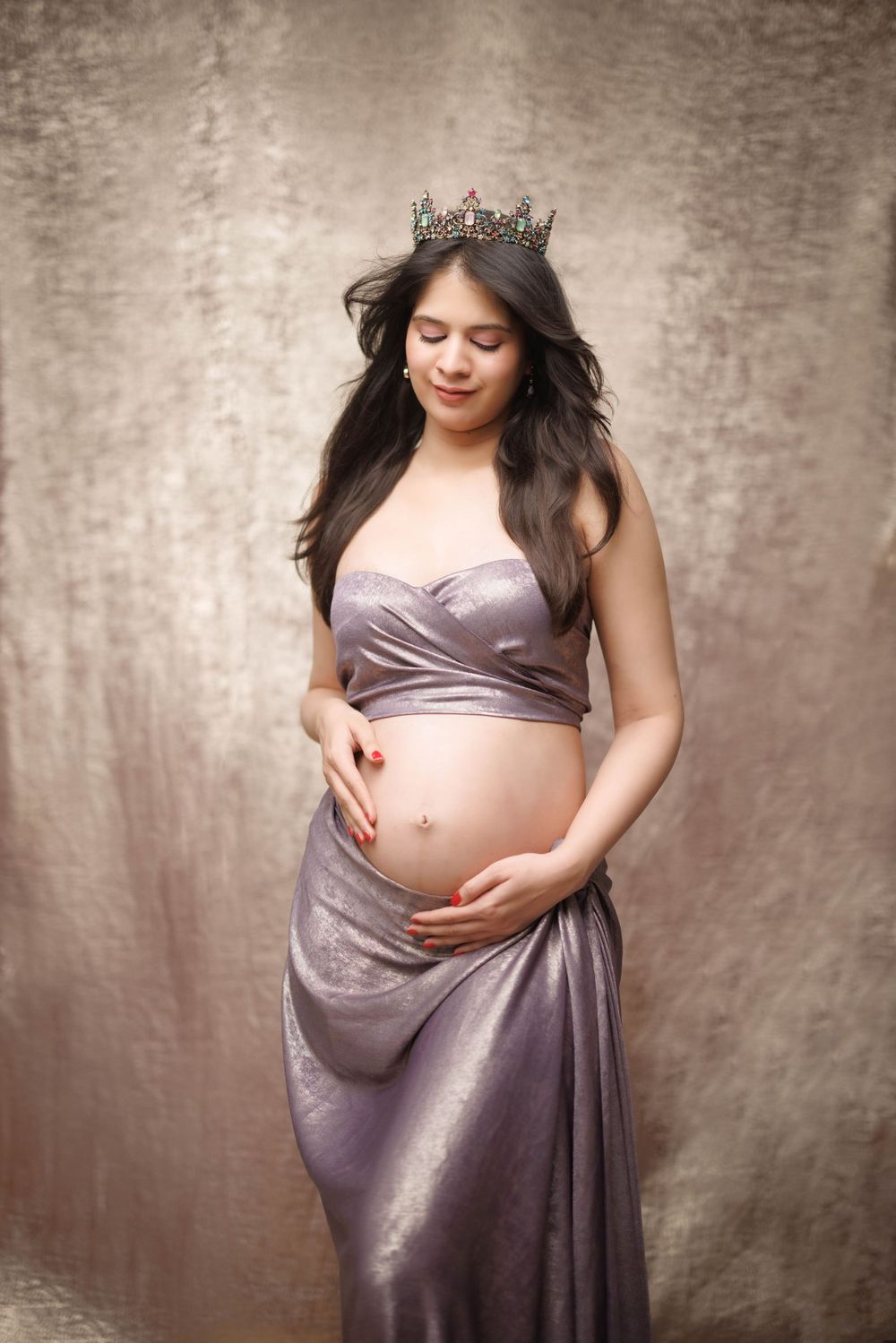 A regal maternity portrait in lavender silk dress with crystal crown against textured backdrop.