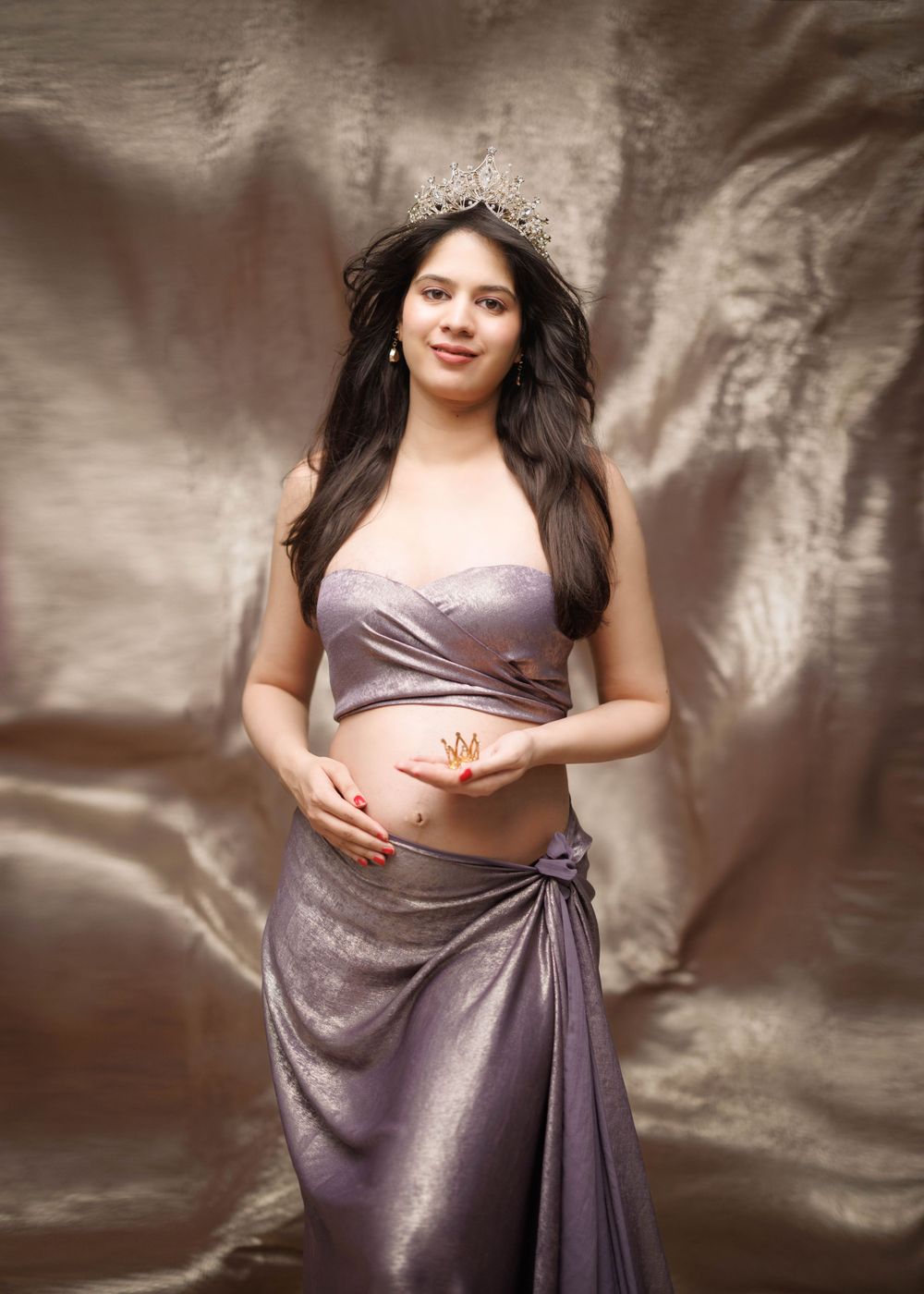 A soft maternity portrait in draped lavender silk with crystal crown against shimmer backdrop.