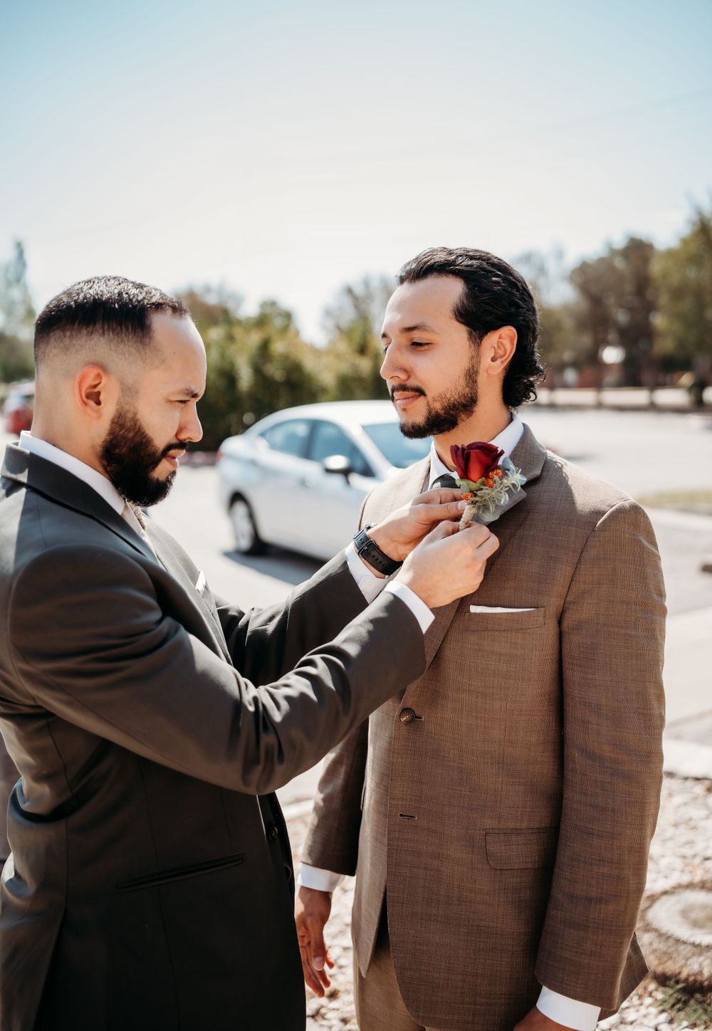 Two men in formal suits adjusting boutonniere outdoors during wedding preparations.