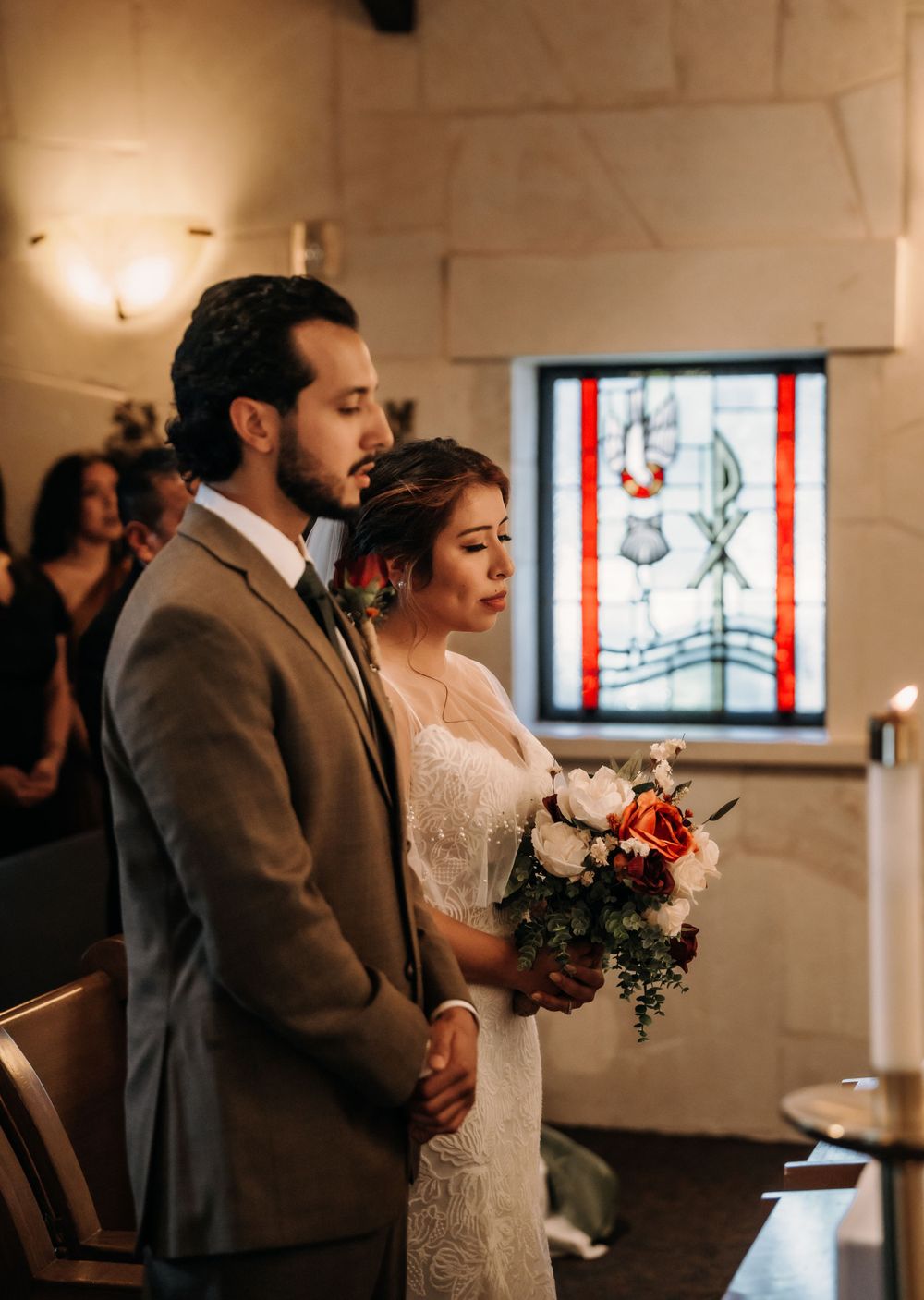 Couple standing together during wedding ceremony in church with stained glass window.