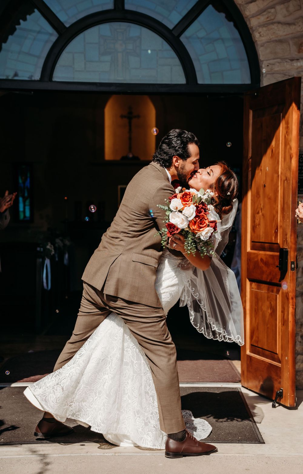 A romantic dip kiss between newlyweds at a church doorway while holding a red and white bouquet.