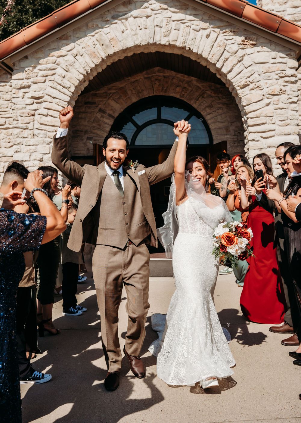 A newly married couple exits a stone church archway while guests cheer during their wedding celebration.