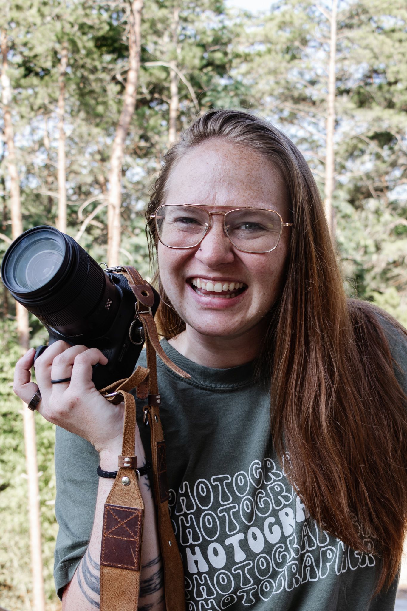 Woman wearing glasses smiling and holding a camera close beside her face with the straps hanging loose. Her shirt says photographer on it.