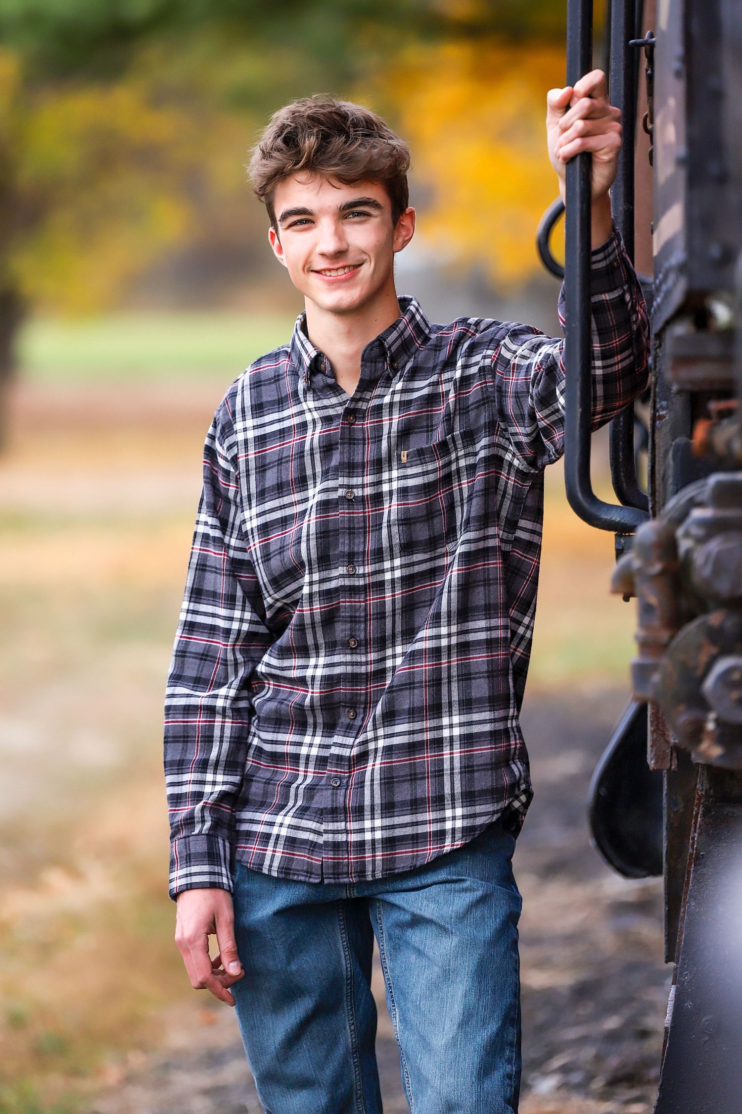 A portrait of someone wearing a plaid flannel shirt and jeans standing next to a railing in autumn.