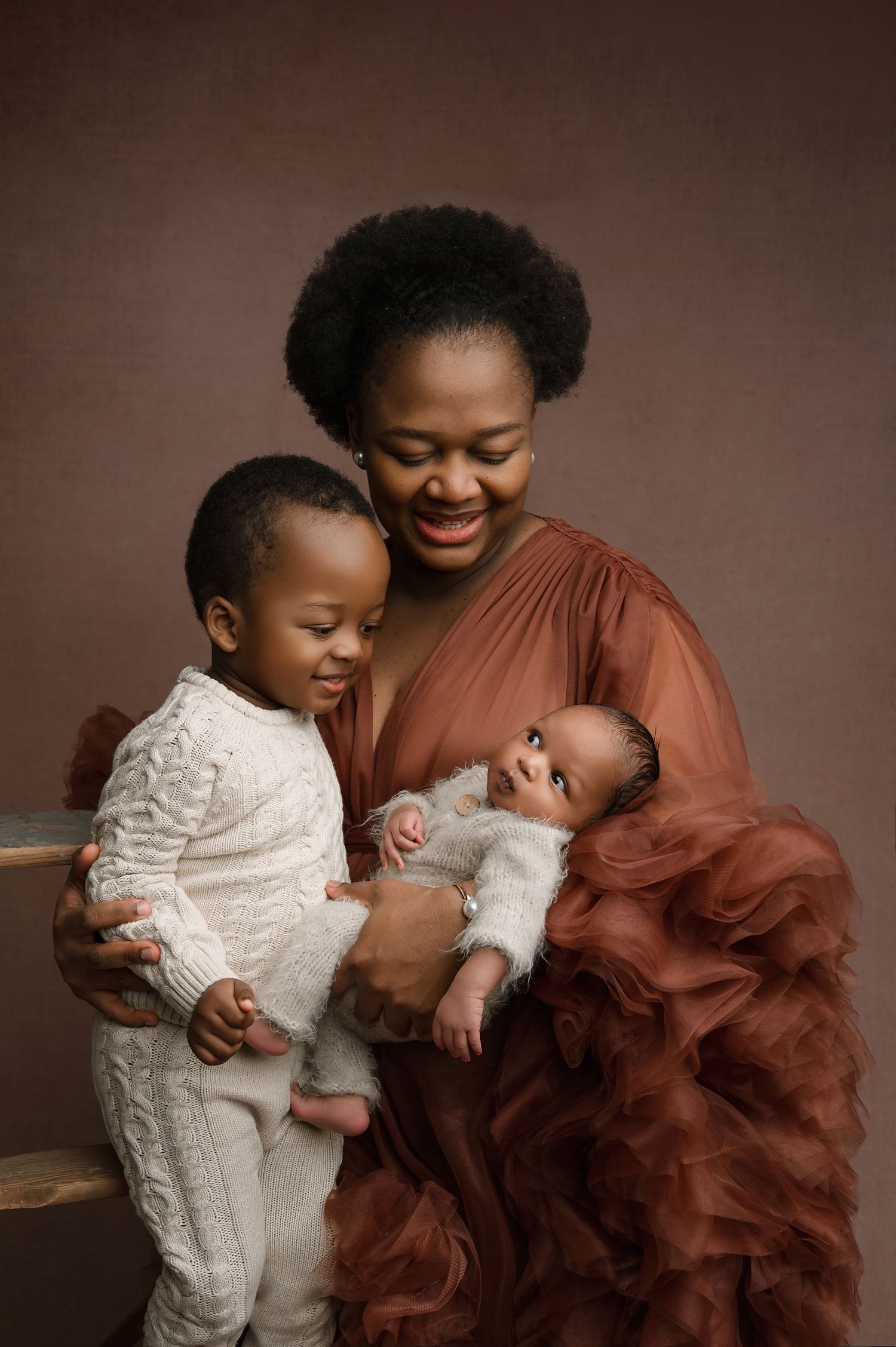 A romantic couple embraces while wearing black and a flowing burgundy gown against a dark gray studio backdrop. newborn photography