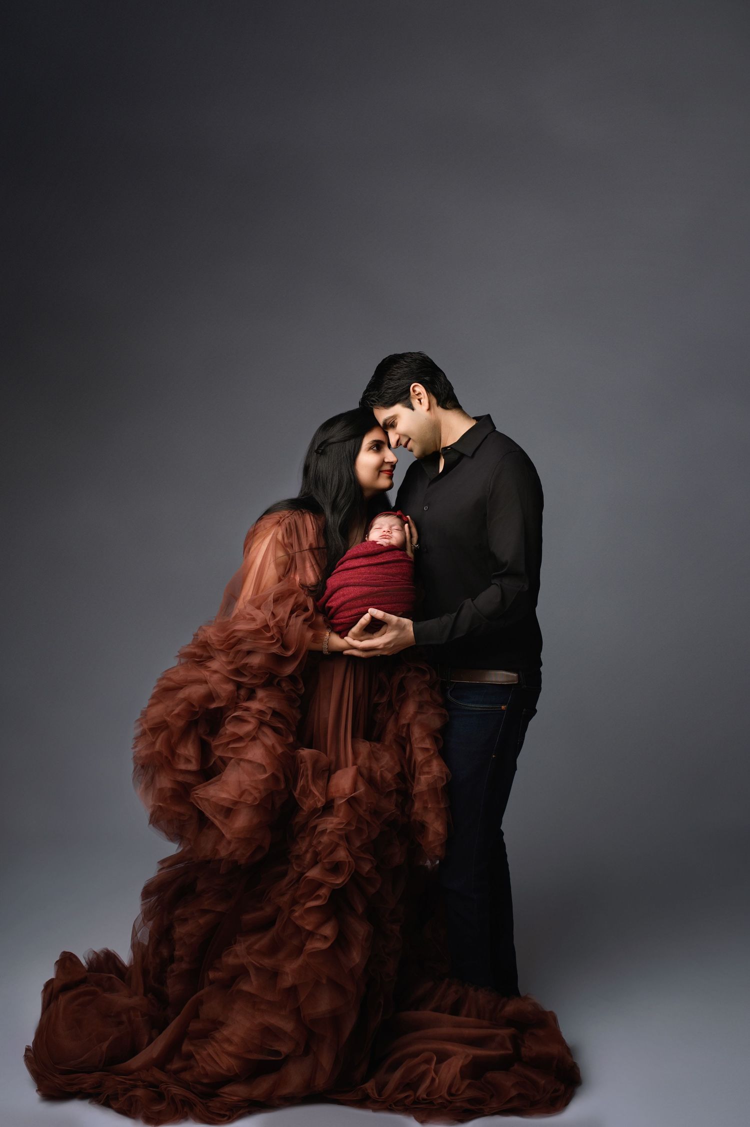 A romantic couple embraces while wearing black and a flowing burgundy gown against a dark gray studio backdrop. newborn photography