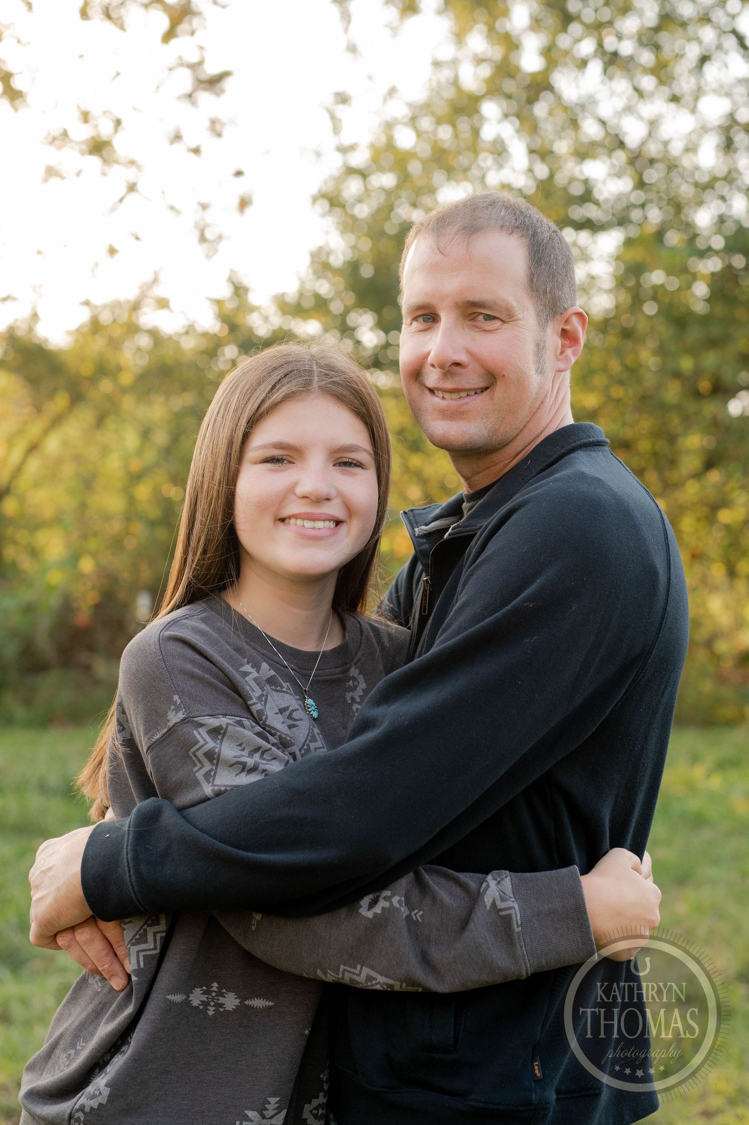 Two people embrace and smile while standing in a sunlit autumn field.