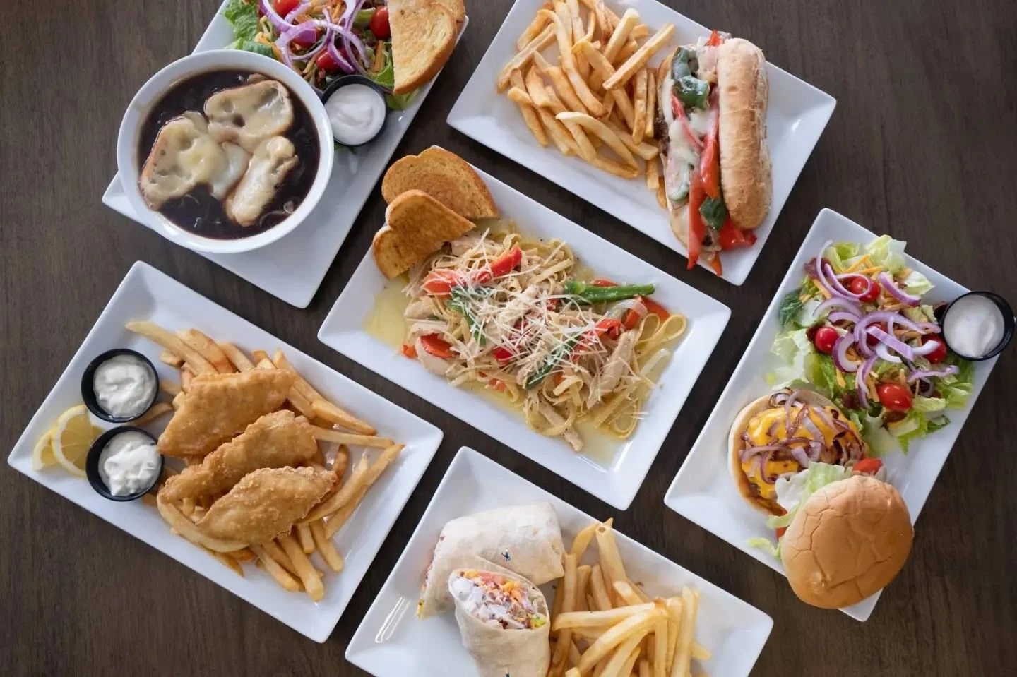 Overhead view of casual restaurant dishes including sandwiches, french fries, and fried seafood with dipping sauces on white plates.