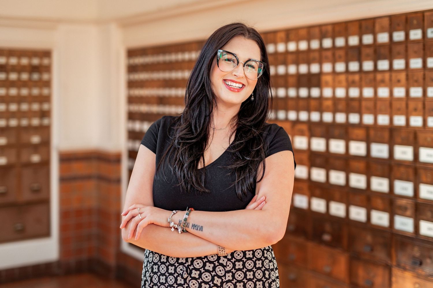 Professional looking woman in black top and patterned skirt stands with arms crossed in office with wooden filing cabinets.
