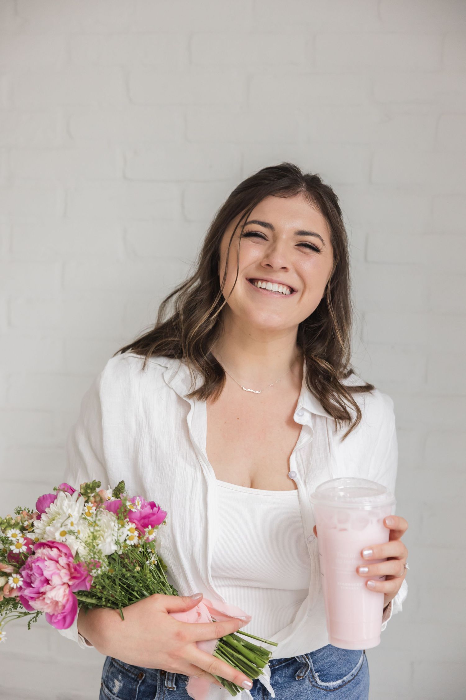 A person in a white top holds a pink smoothie and colorful bouquet of flowers against a light background.