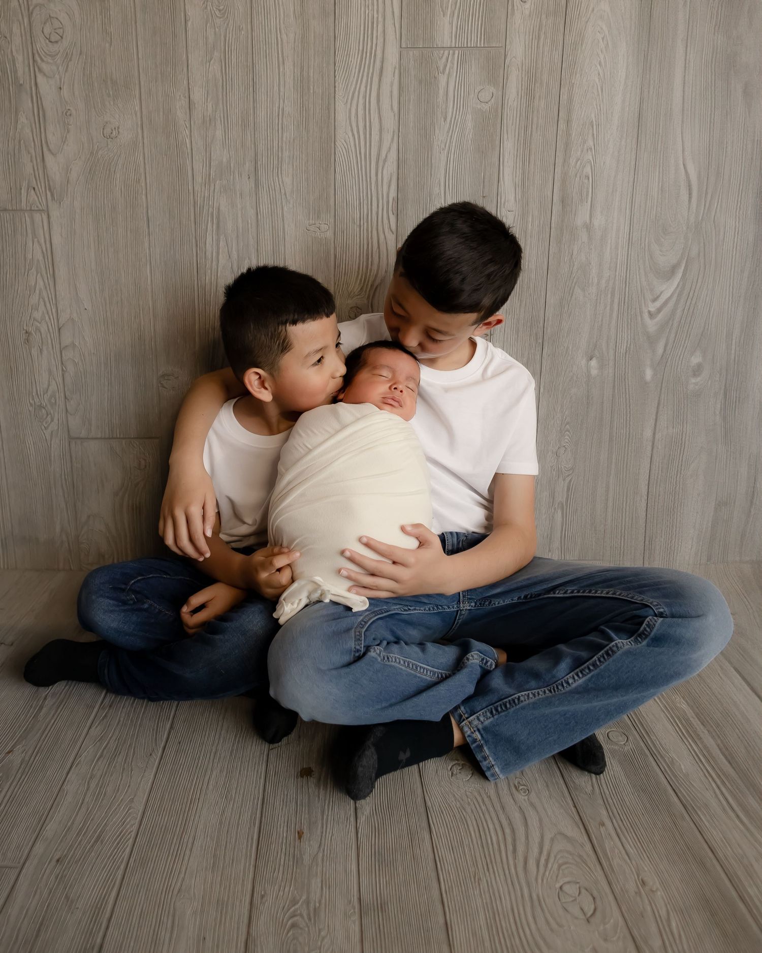 Two siblings in jeans and white shirts sit on a wood-paneled floor while gently holding a swaddled baby.