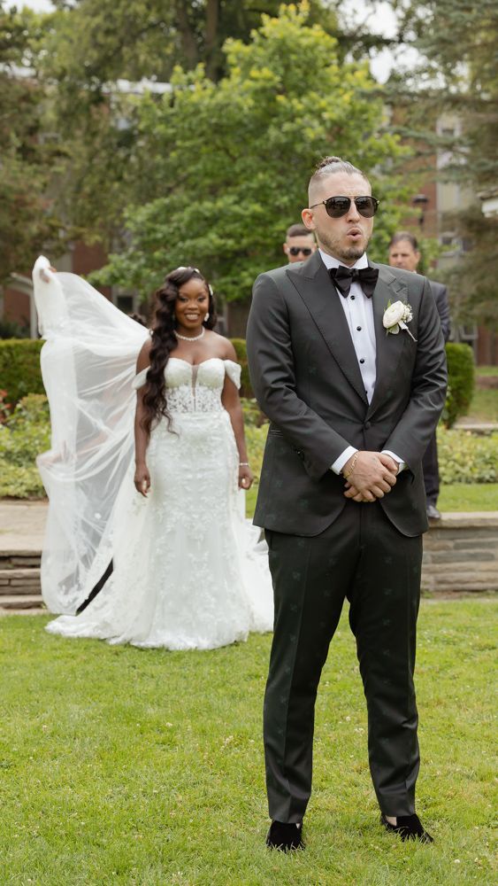A wedding couple poses in a garden, with the bride in a white lace gown and the groom wearing a formal black tuxedo and sunglasses.