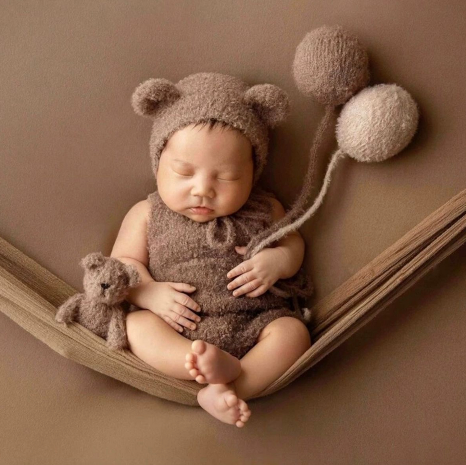 Baby wearing a soft brown teddy bear outfit sleeping on brown backdrop.