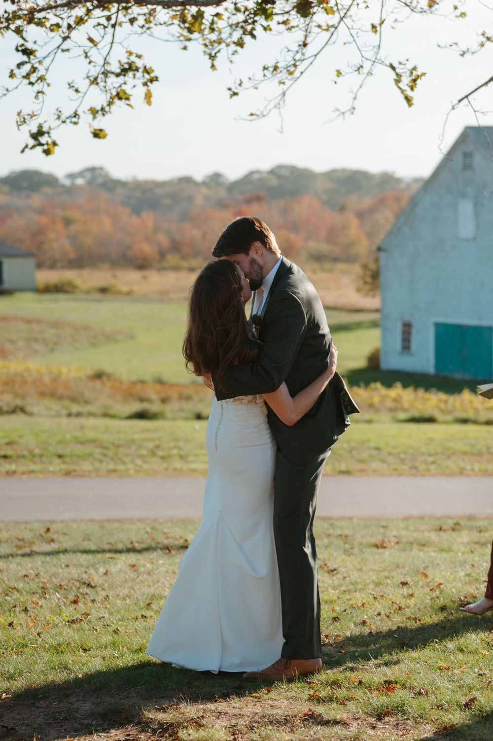 Couple embracing in an autumn field with white farmhouse buildings visible in the background.