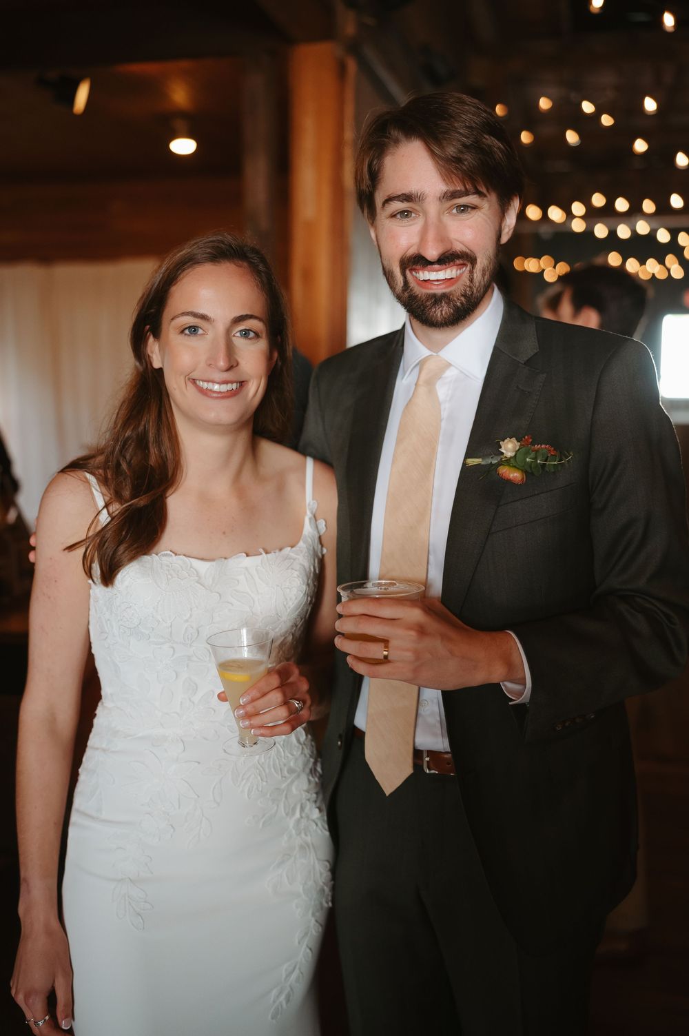 A couple shares drinks and smiles at their wedding reception under warm string lights.