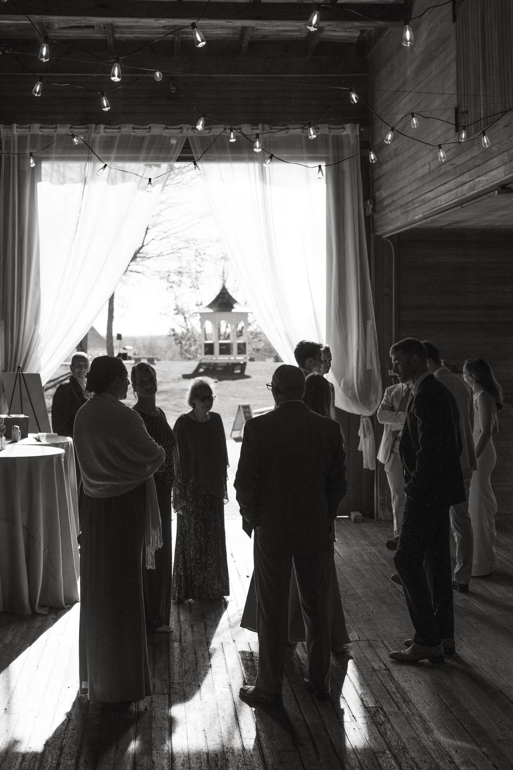 Silhouettes of wedding guests mingle in a bright venue with sunlight streaming through white curtained windows.