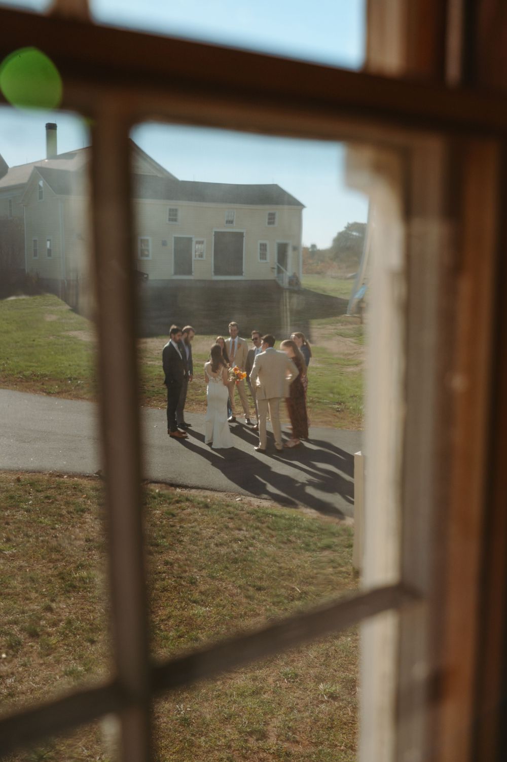 View through a window of wedding guests gathering outside a rural building.