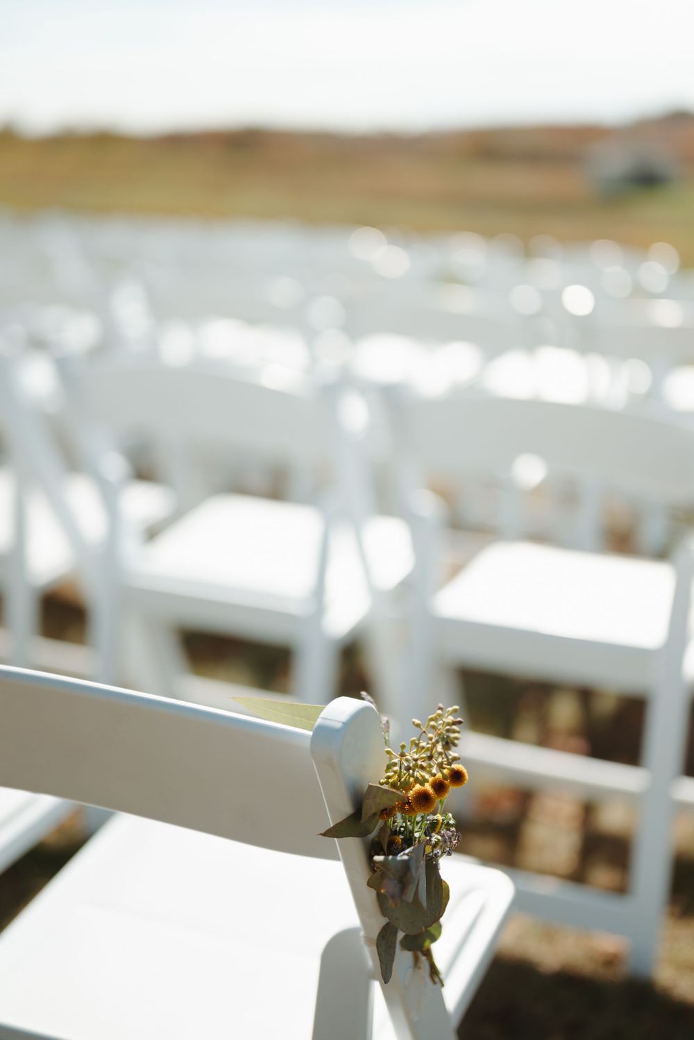 White folding chairs adorned with small floral arrangements set up for an outdoor wedding ceremony.